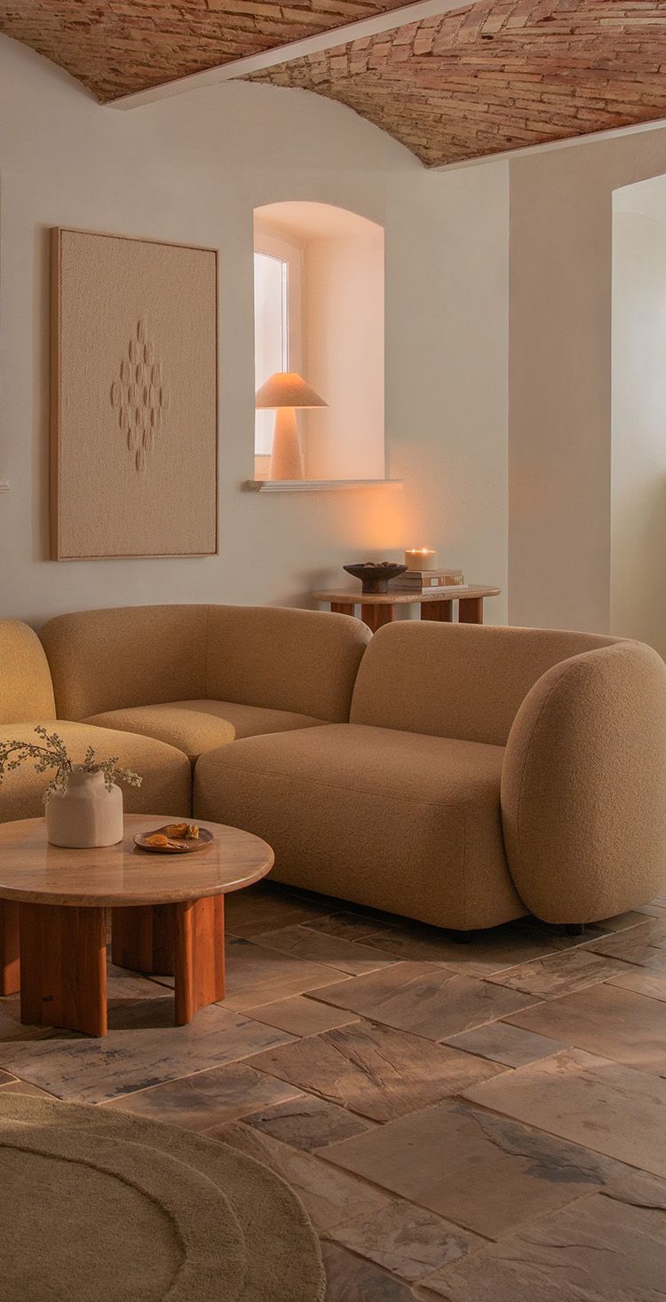 Canapé d’angle Micadoni Kate en revêtement beige chaud, mis en scène dans un salon chaleureux avec éclairage d’ambiance doux et table basse ronde en bois.