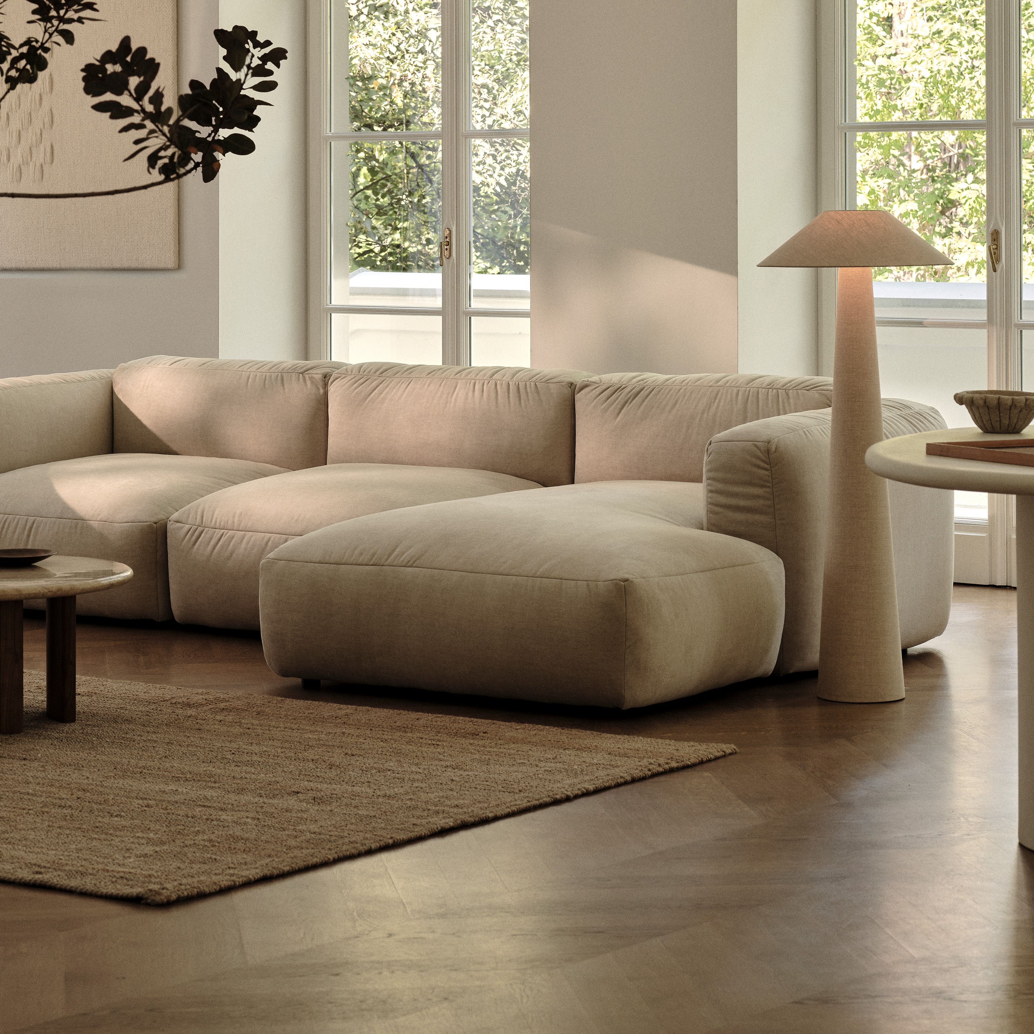 Canapé modulaire Micadoni Martina en tissu beige avec grande méridienne, dans un salon lumineux au sol en bois, table basse ronde et lampe sur pied conique.