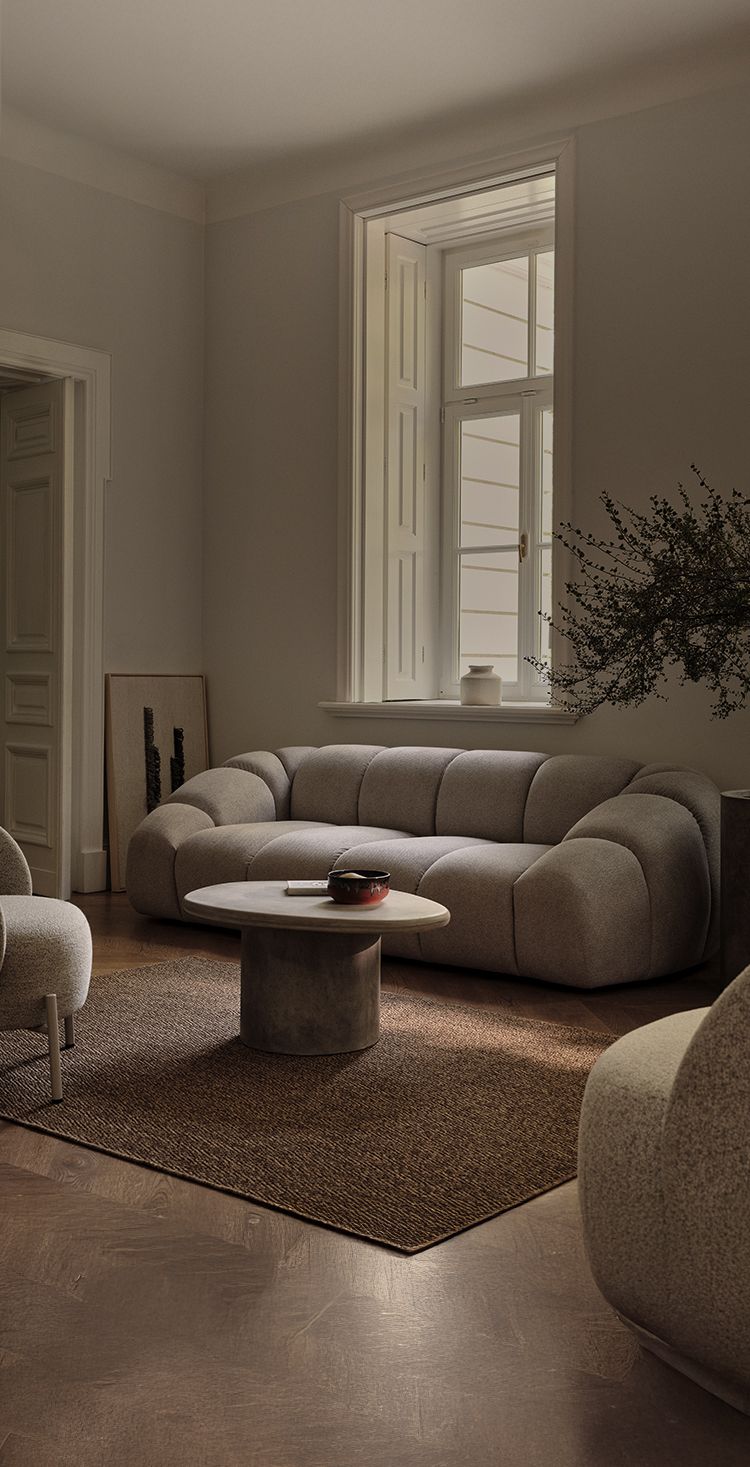 Canapé Micadoni Diana aux volumes arrondis et coutures cannelées en tissu gris, associé à la table basse Daria aspect béton, dans un salon doux avec tapis texturé.