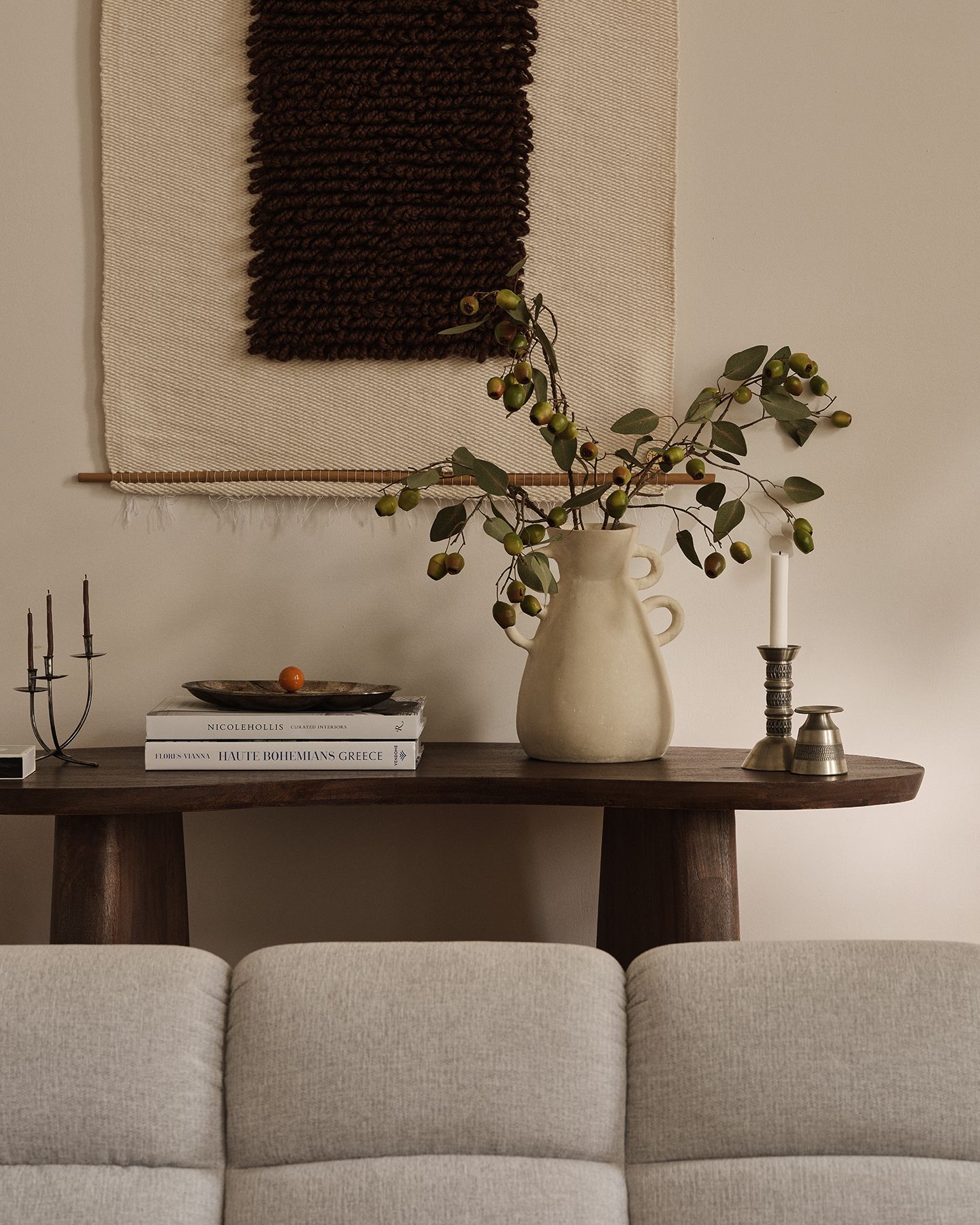 Scène salon Micadoni avec console en bois foncé, pichet en céramique et branches, livres et bougie, tenture murale textile et dossier d’un canapé gris clair.