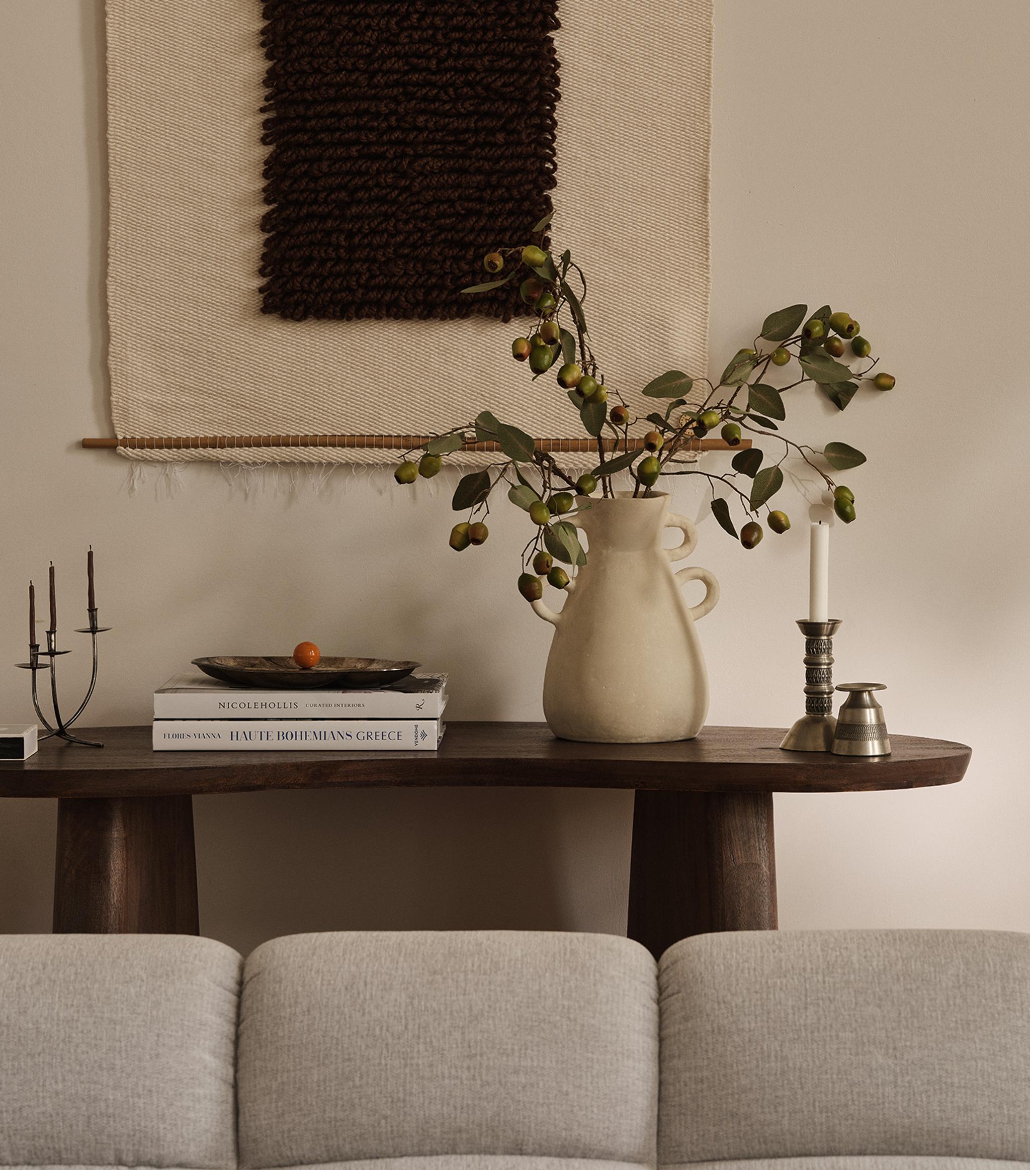 Scène salon Micadoni avec console en bois foncé, pichet en céramique et branches, livres et bougie, tenture murale textile et dossier d’un canapé gris clair.