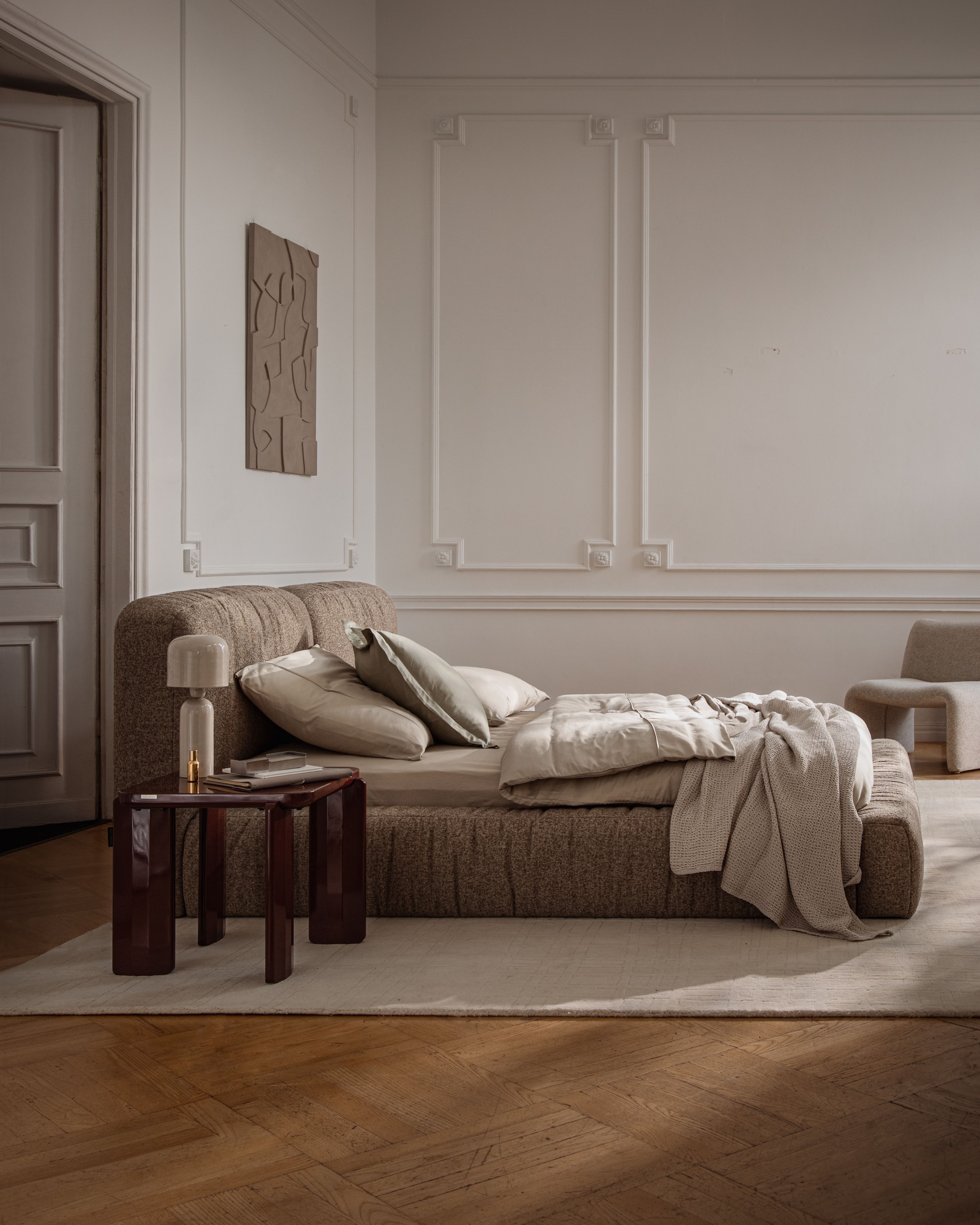 Lit Micadoni Martina en tissu avec tête de lit basse, table d’appoint et linge de lit neutre dans un intérieur classique.