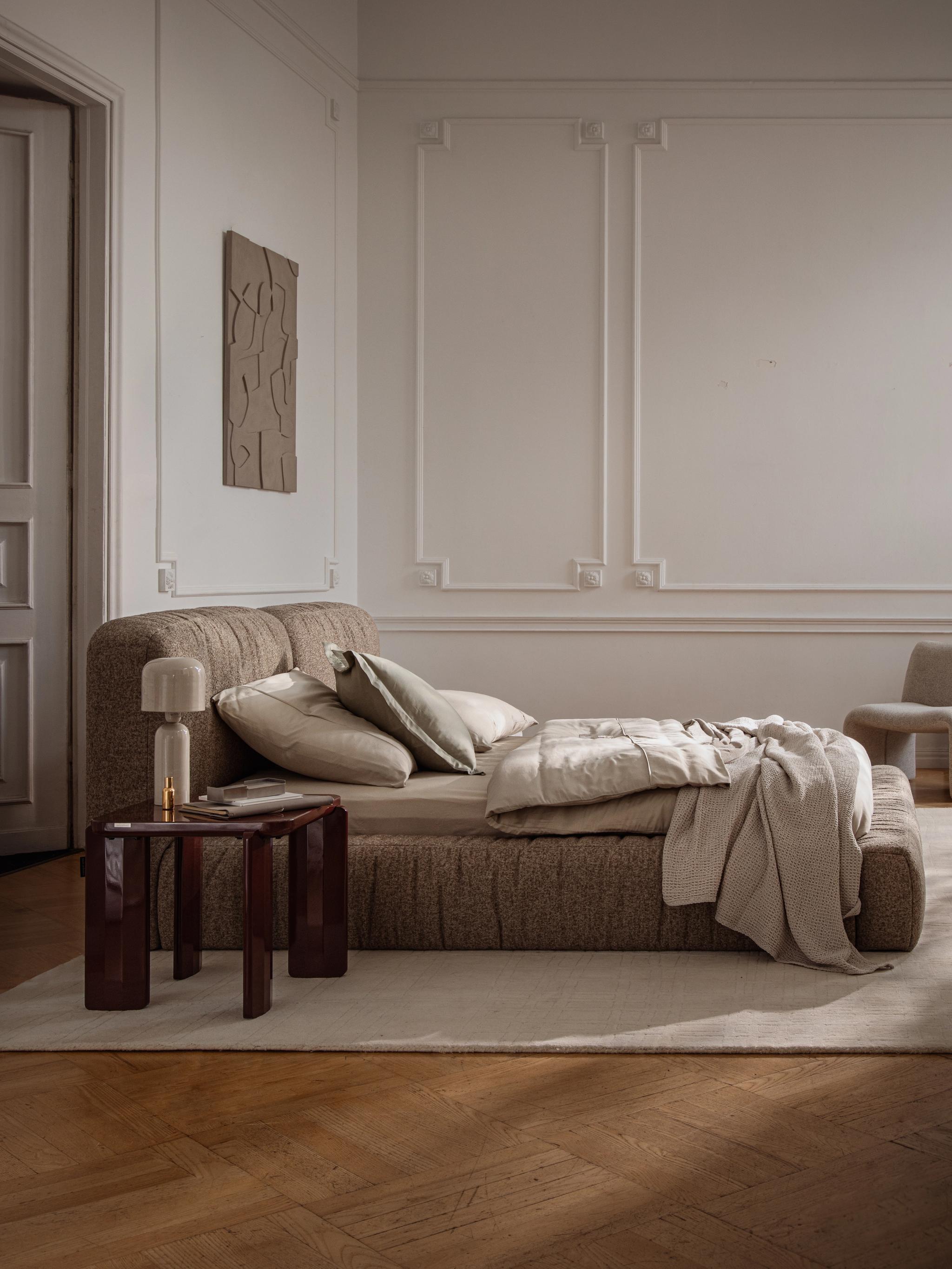 Lit Micadoni Martina en tissu avec tête de lit basse, table d’appoint et linge de lit neutre dans un intérieur classique.