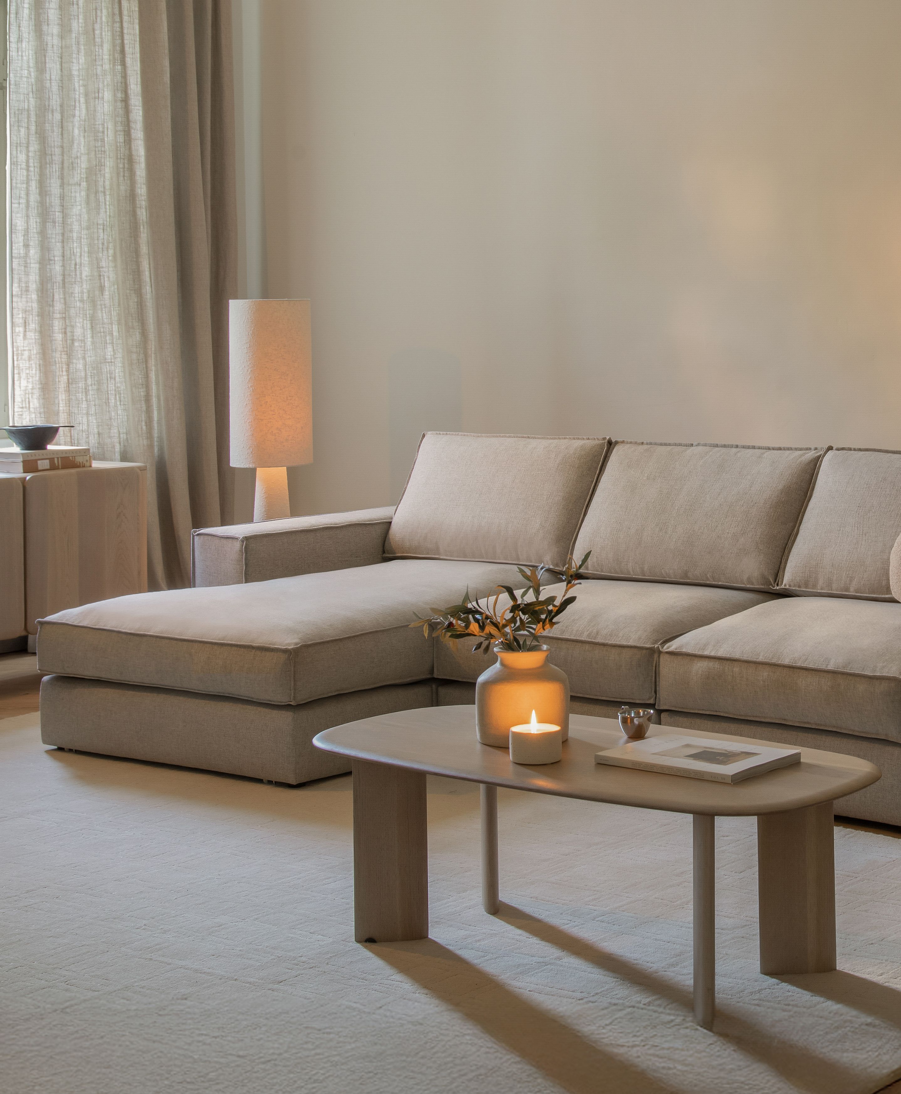 Canapé d’angle modulaire Pola Micadoni en tissu beige avec méridienne à gauche, salon apaisé avec table basse, bougie et lampadaire.