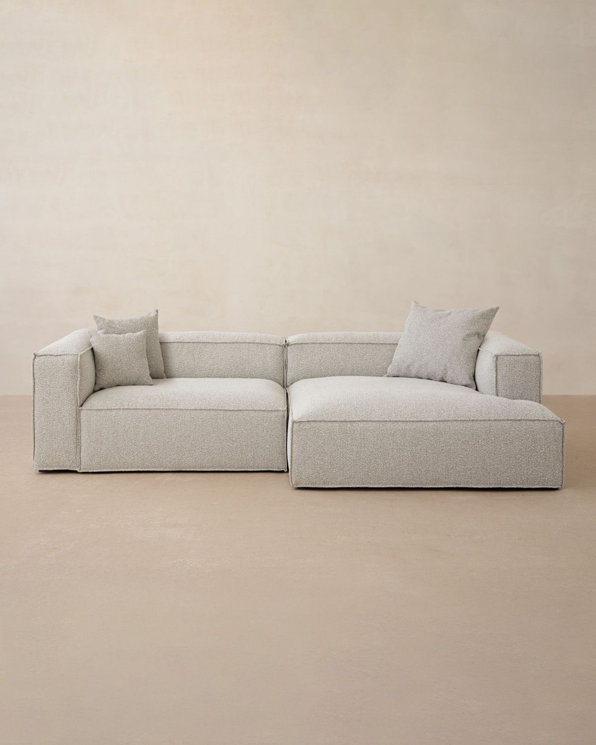 Canapé d’angle Micadoni Nuria en bouclé gris clair avec méridienne à droite et accoudoirs larges, décor neutre.