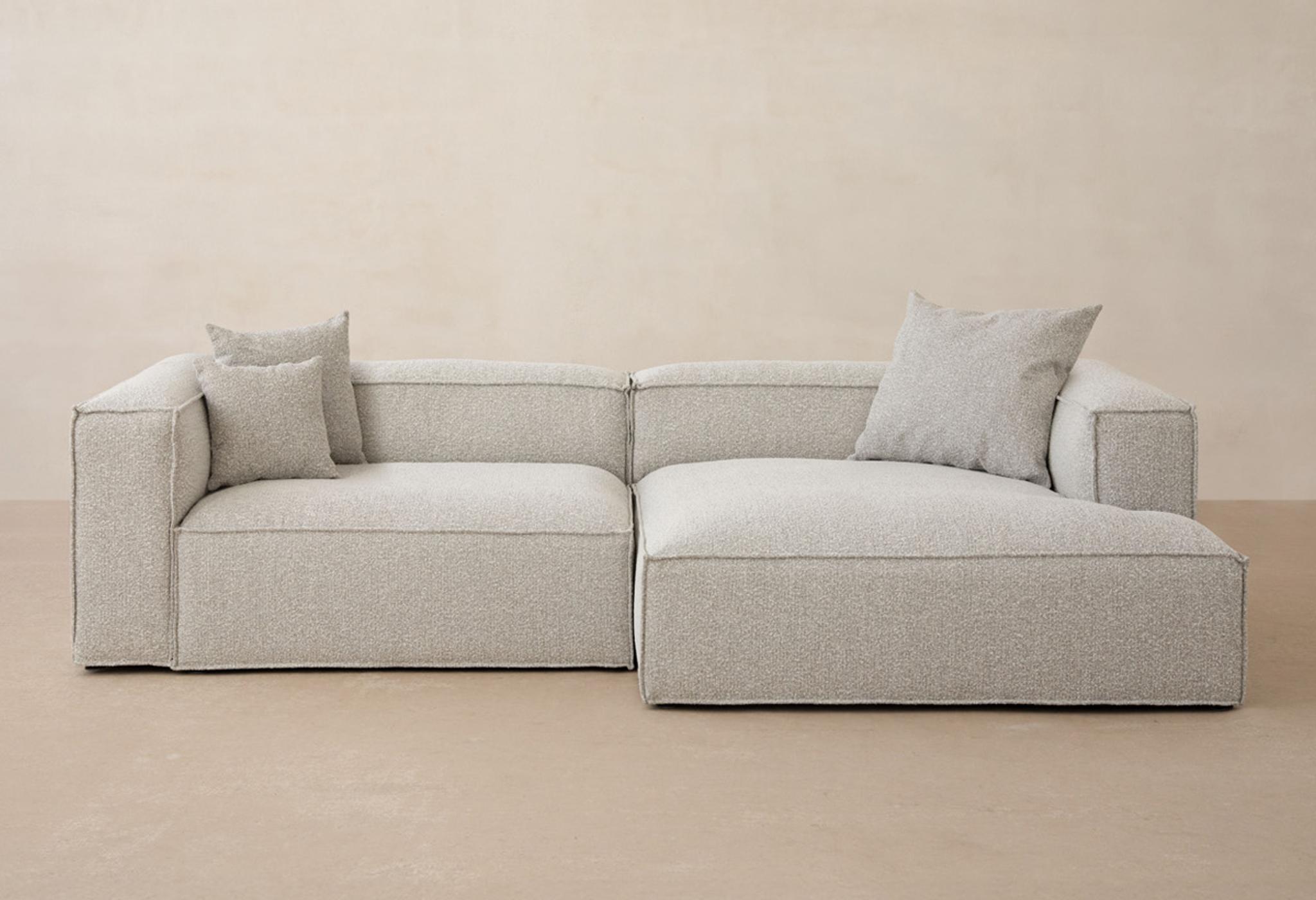 Canapé d’angle Micadoni Nuria en bouclé gris clair avec méridienne à droite et accoudoirs larges, décor neutre.