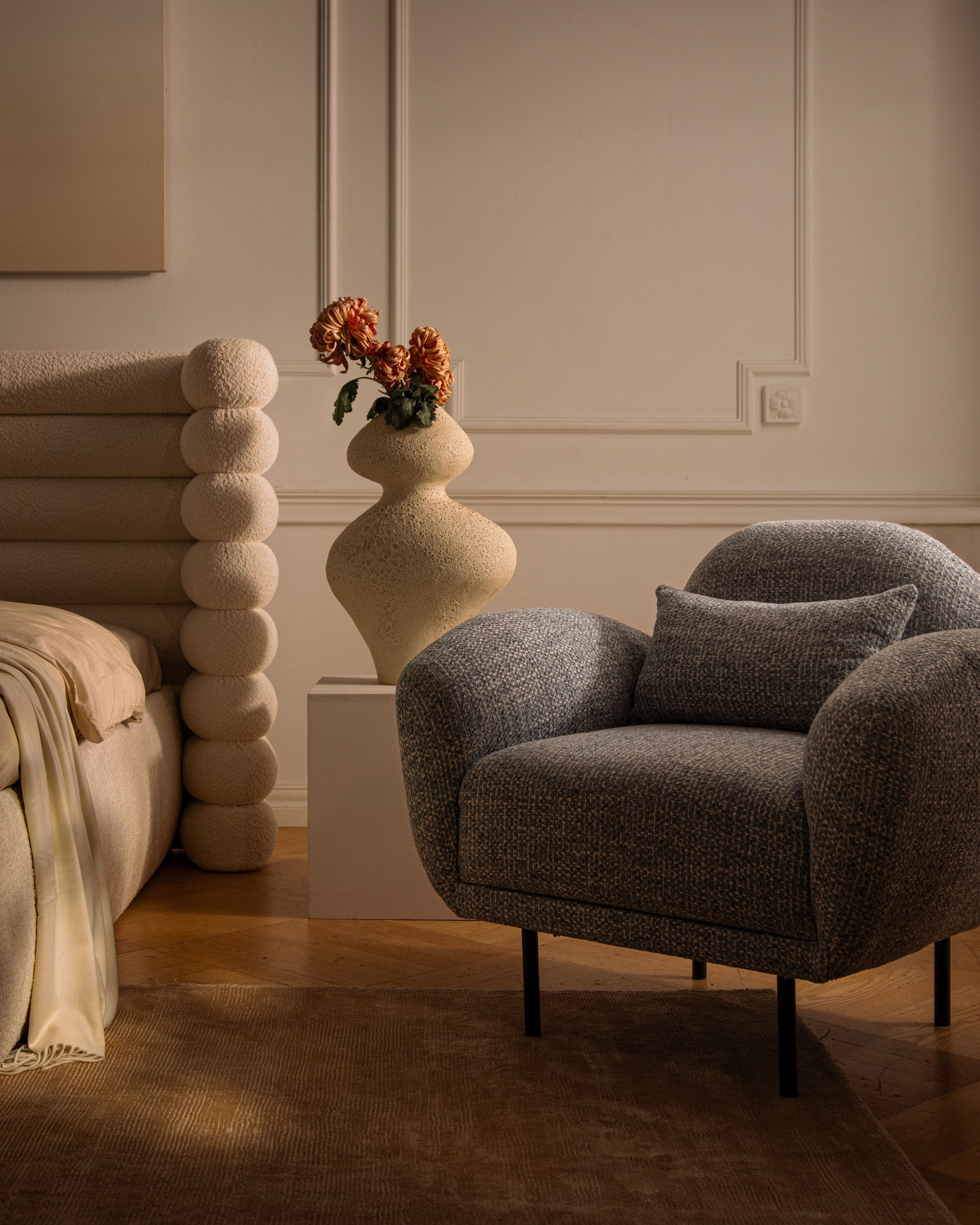 Lit Micadoni avec tête de lit capitonnée arrondie et fauteuil gris texturé dans une chambre chaleureuse.