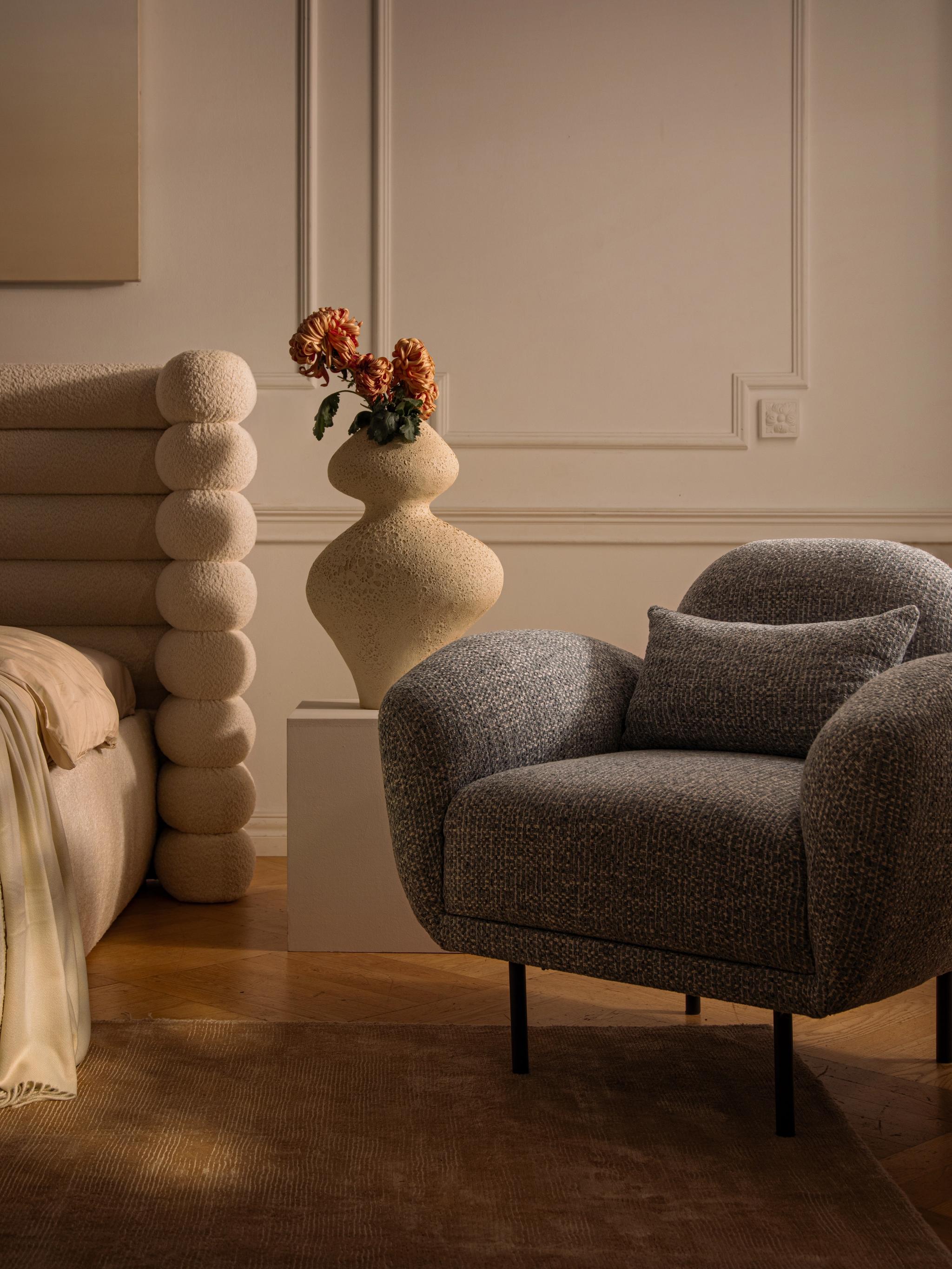 Lit Micadoni avec tête de lit capitonnée arrondie et fauteuil gris texturé dans une chambre chaleureuse.