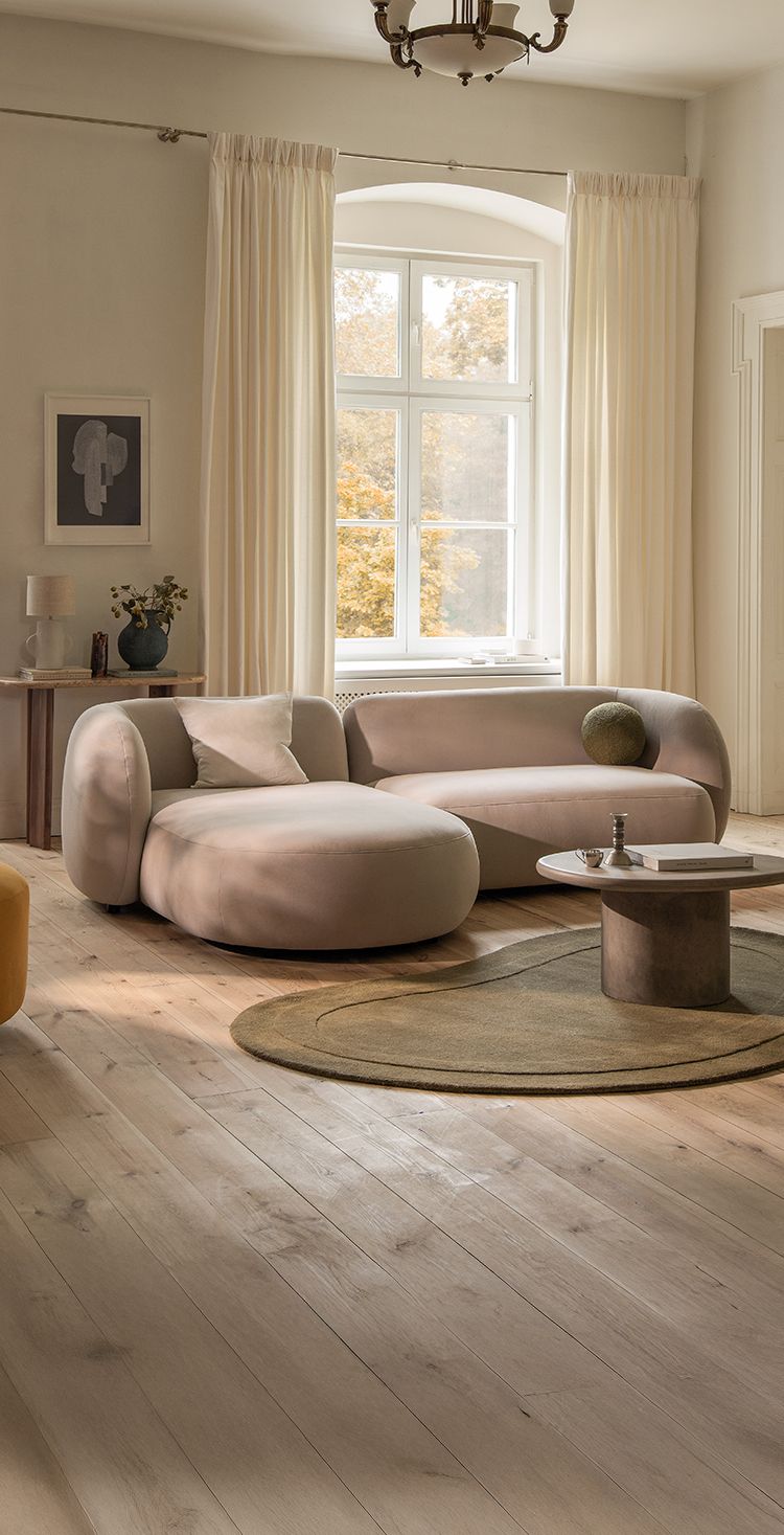 Micadoni Kate corner sofa in a soft beige fabric with rounded arms and a chaise on the left, styled in a bright living room with a round pedestal coffee table and an olive oval rug.