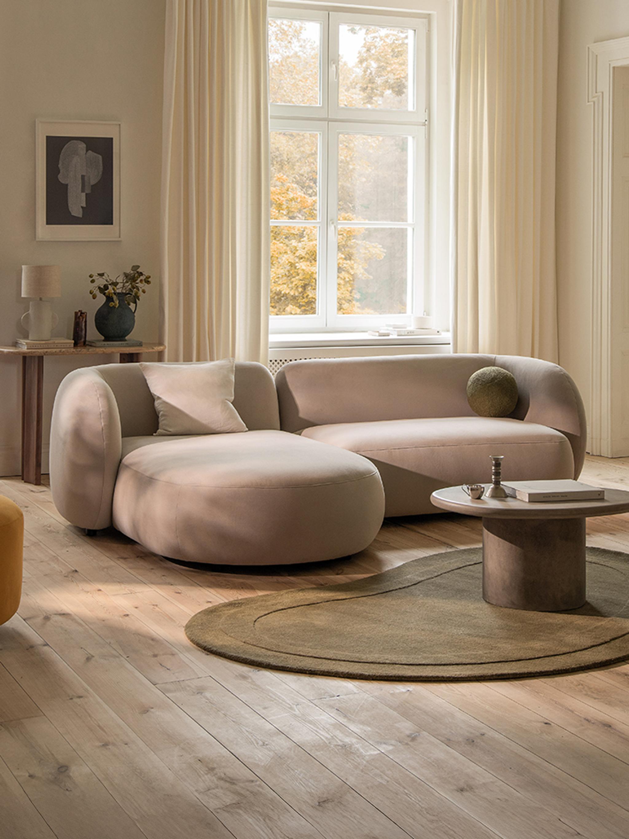 Micadoni Kate corner sofa in a soft beige fabric with rounded arms and a chaise on the left, styled in a bright living room with a round pedestal coffee table and an olive oval rug.