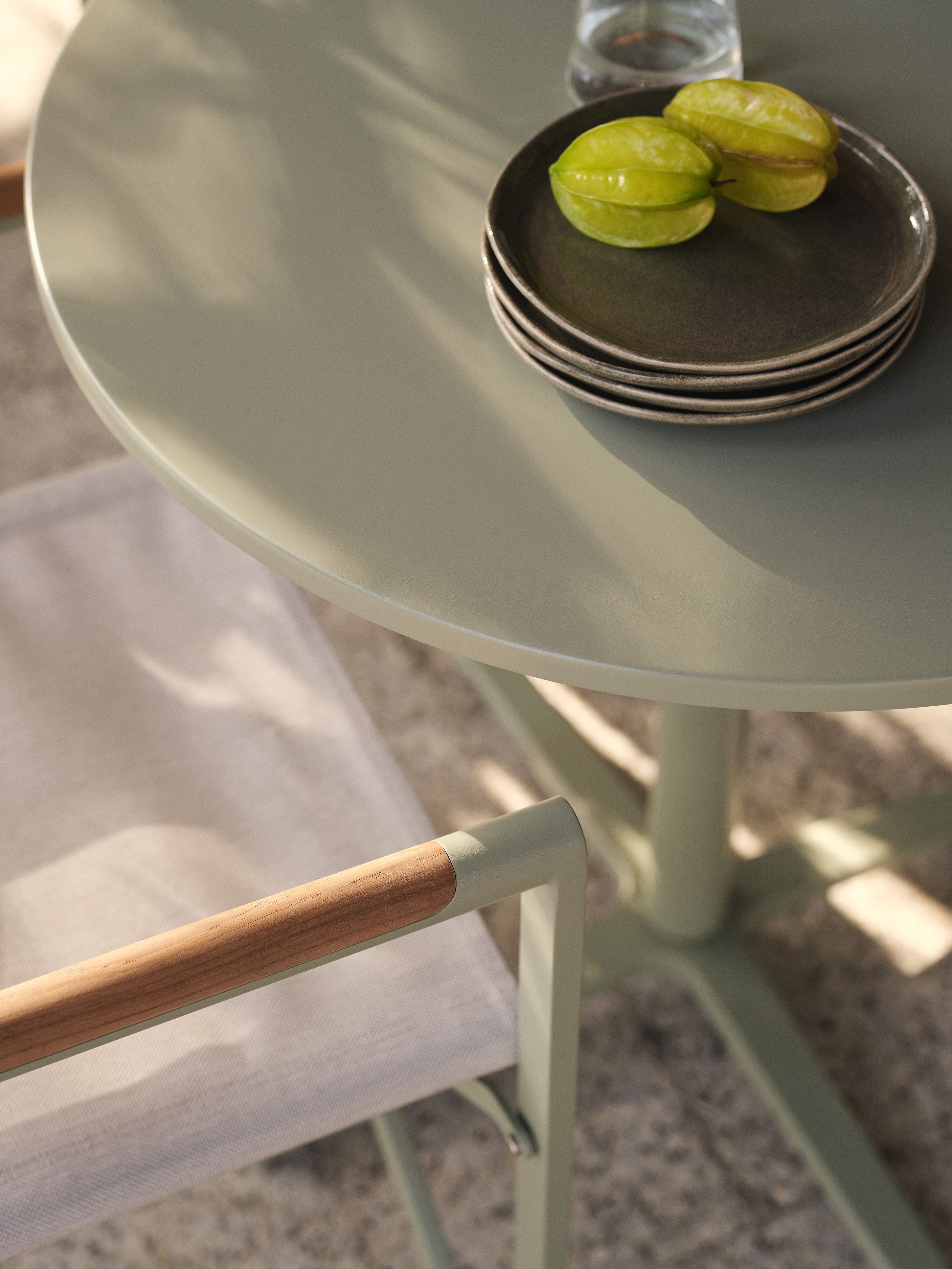 Gros plan sur une table d’extérieur Micadoni ronde vert sauge, plateau brillant, assiettes empilées et carambole.