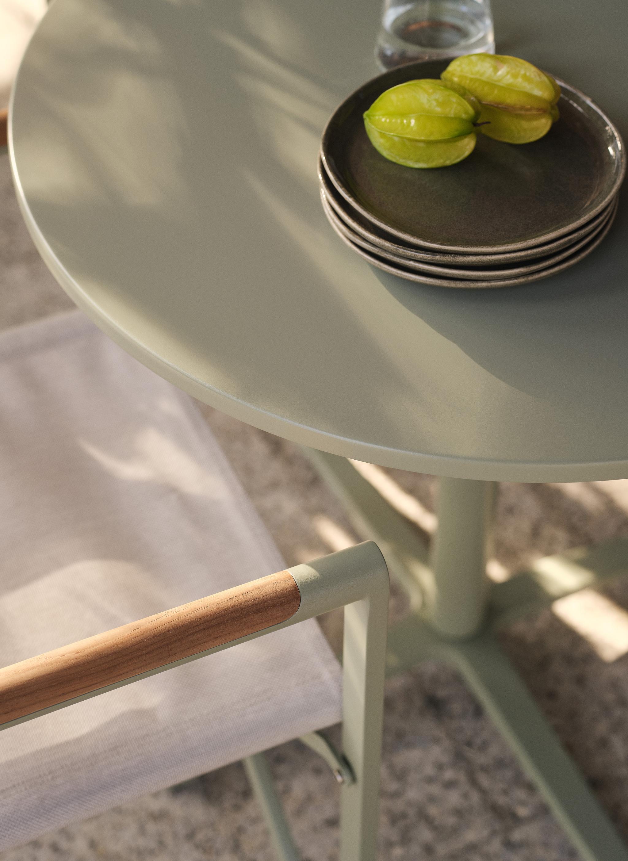 Gros plan sur une table d’extérieur Micadoni ronde vert sauge, plateau brillant, assiettes empilées et carambole.