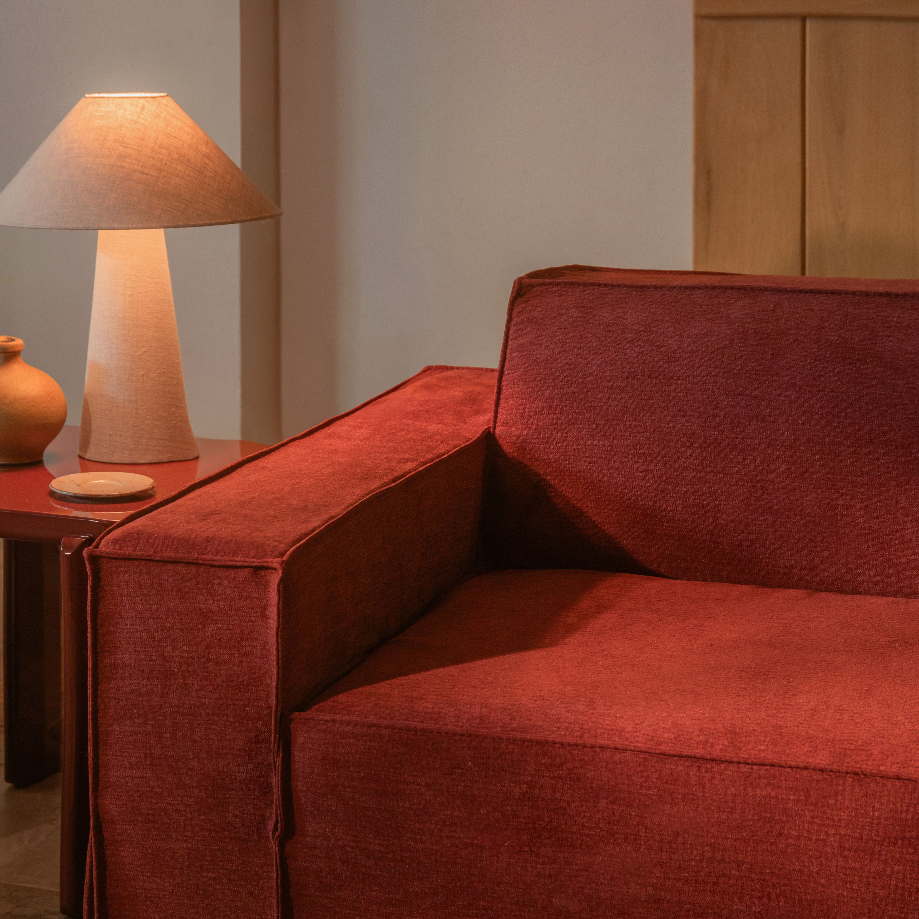 Gros plan sur le canapé Jodie de Micadoni en tissu rouge profond avec accoudoir droit, à côté d’une lampe beige.