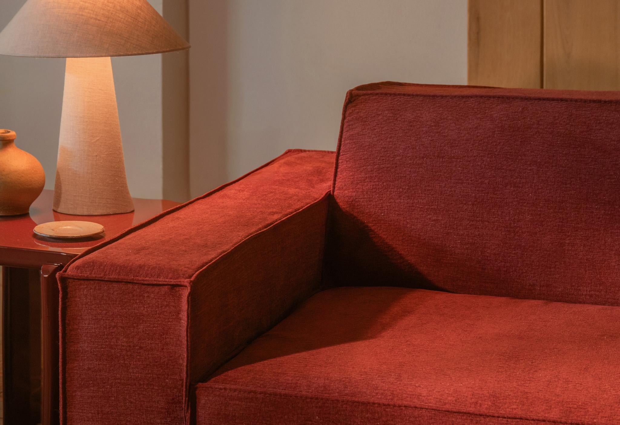 Gros plan sur le canapé Jodie de Micadoni en tissu rouge profond avec accoudoir droit, à côté d’une lampe beige.