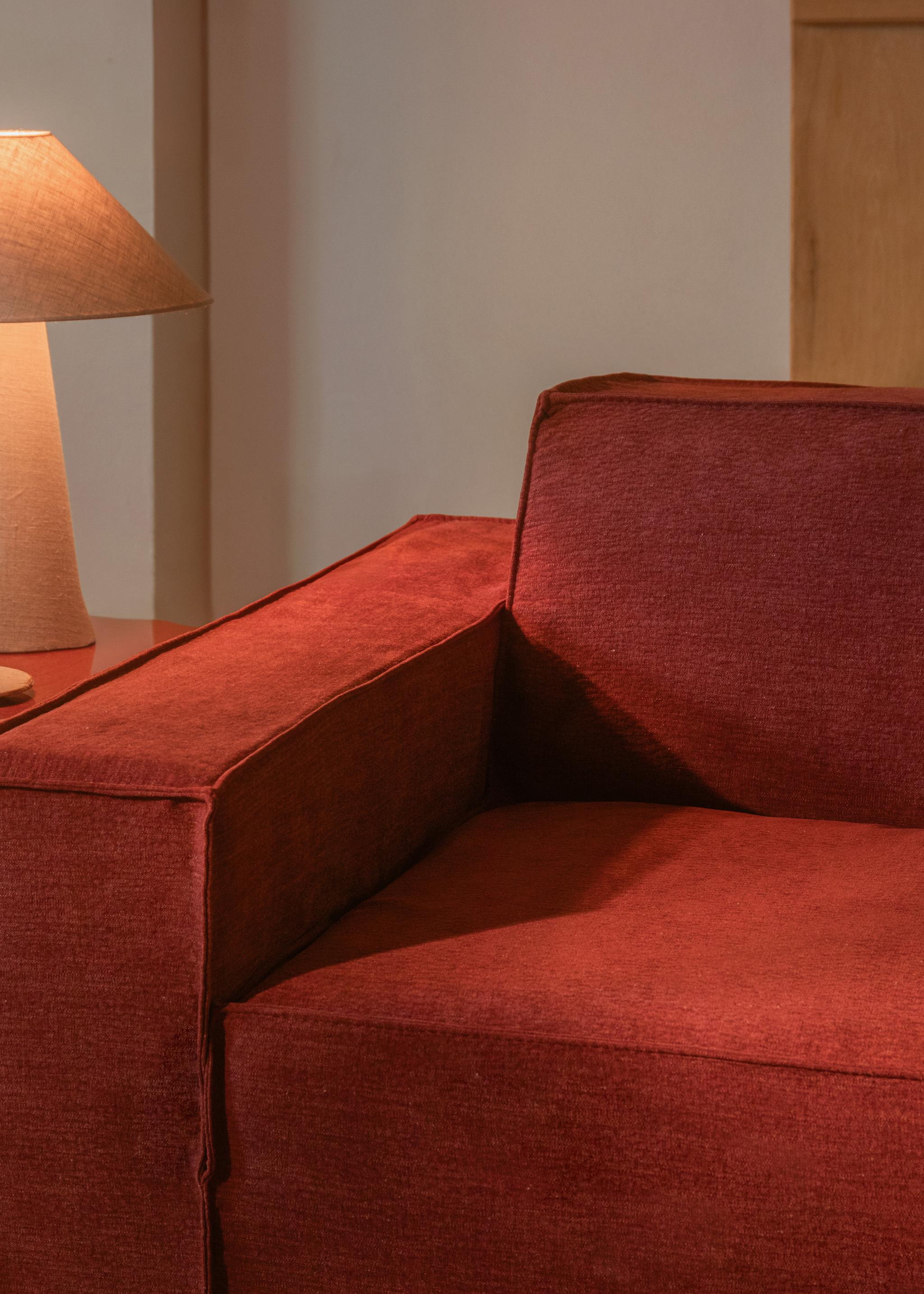 Gros plan sur le canapé Jodie de Micadoni en tissu rouge profond avec accoudoir droit, à côté d’une lampe beige.