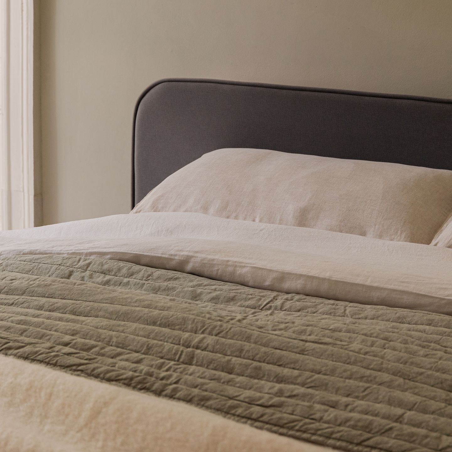 Gros plan sur un lit Micadoni avec tête de lit arrondie capitonnée gris foncé, draps en lin beige et couvre-lit olive.