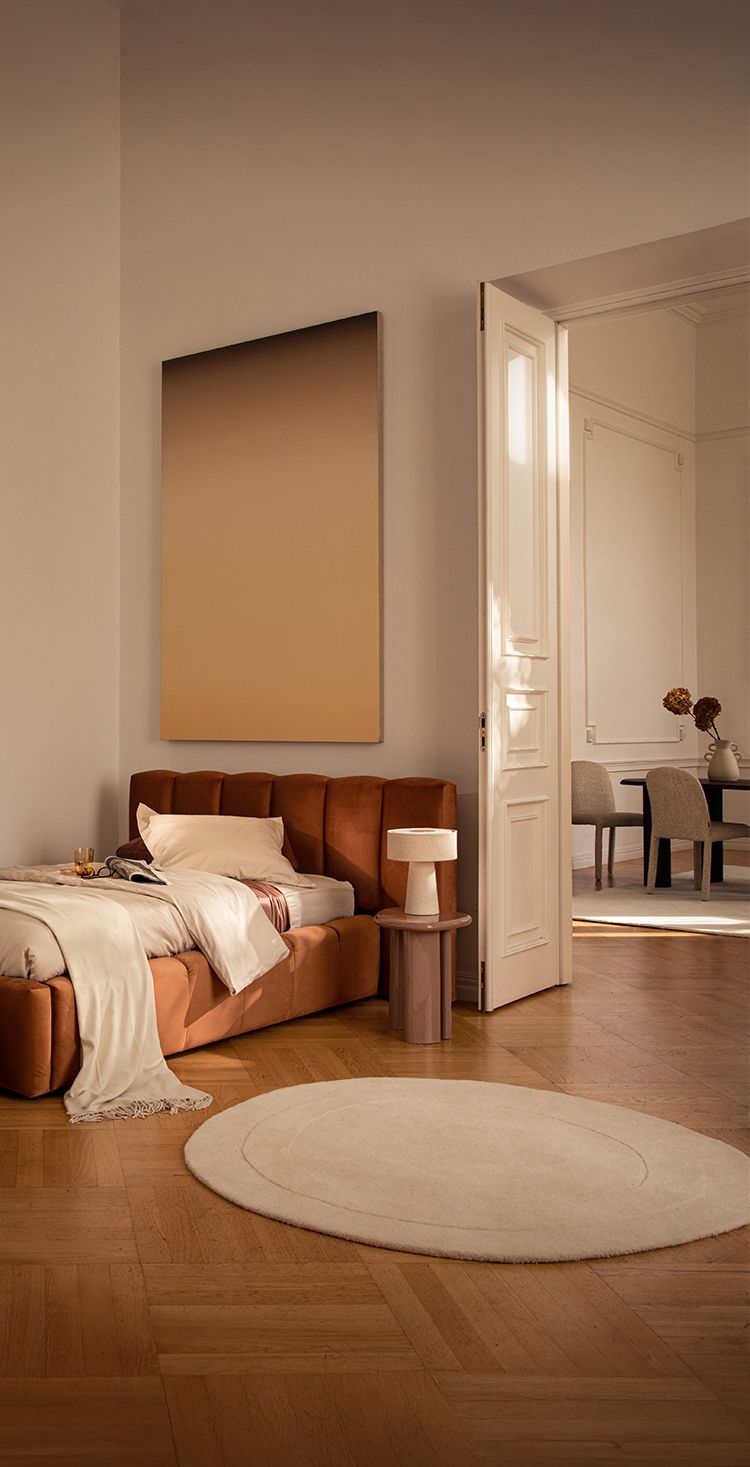 Lit Micadoni Lupine en revêtement rouille chaleureux avec tête de lit à coutures verticales, mis en scène dans une chambre aux tons doux avec tapis rond crème, table d’appoint et lampe de table.