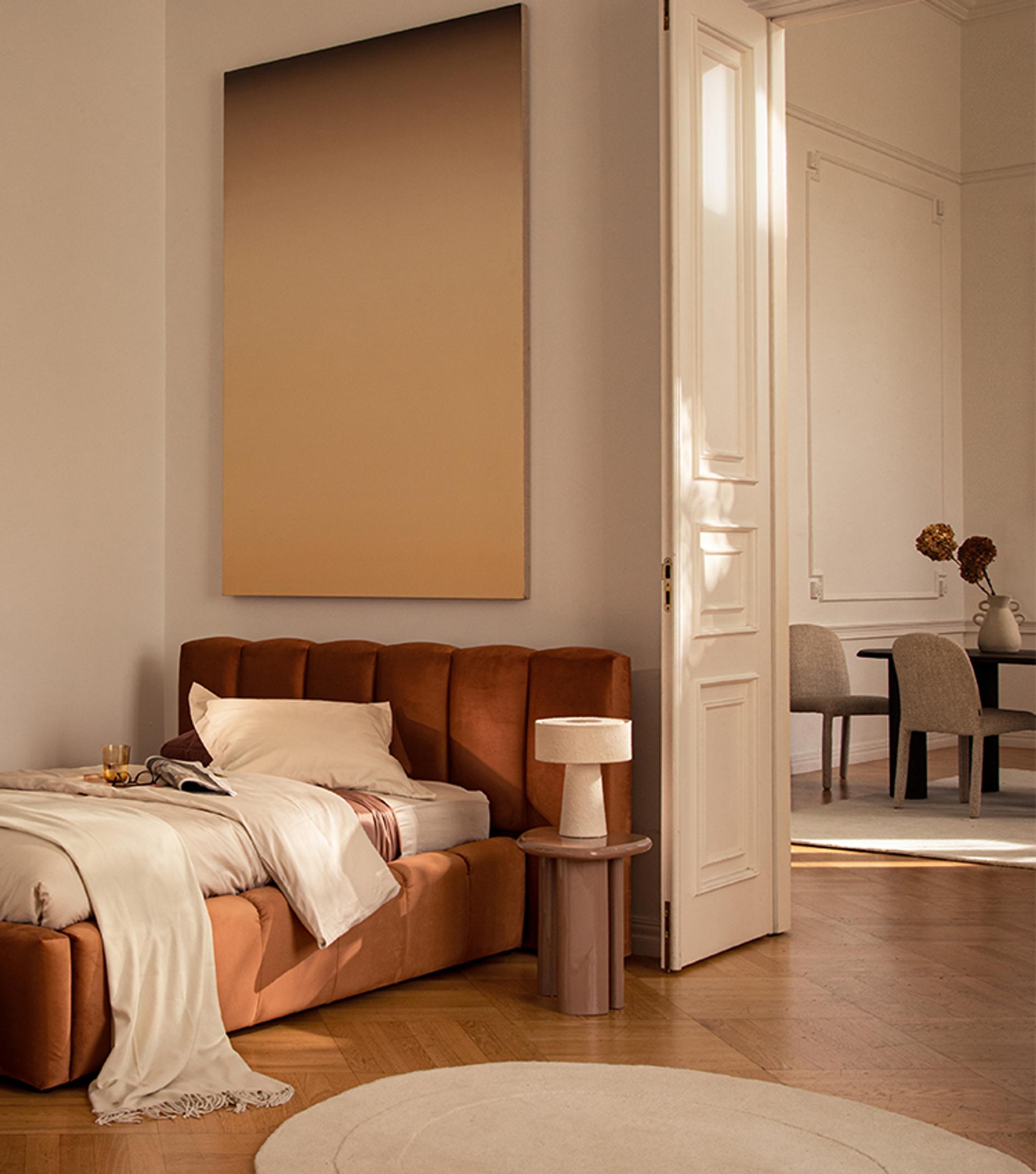 Lit Micadoni Lupine en revêtement rouille chaleureux avec tête de lit à coutures verticales, mis en scène dans une chambre aux tons doux avec tapis rond crème, table d’appoint et lampe de table.