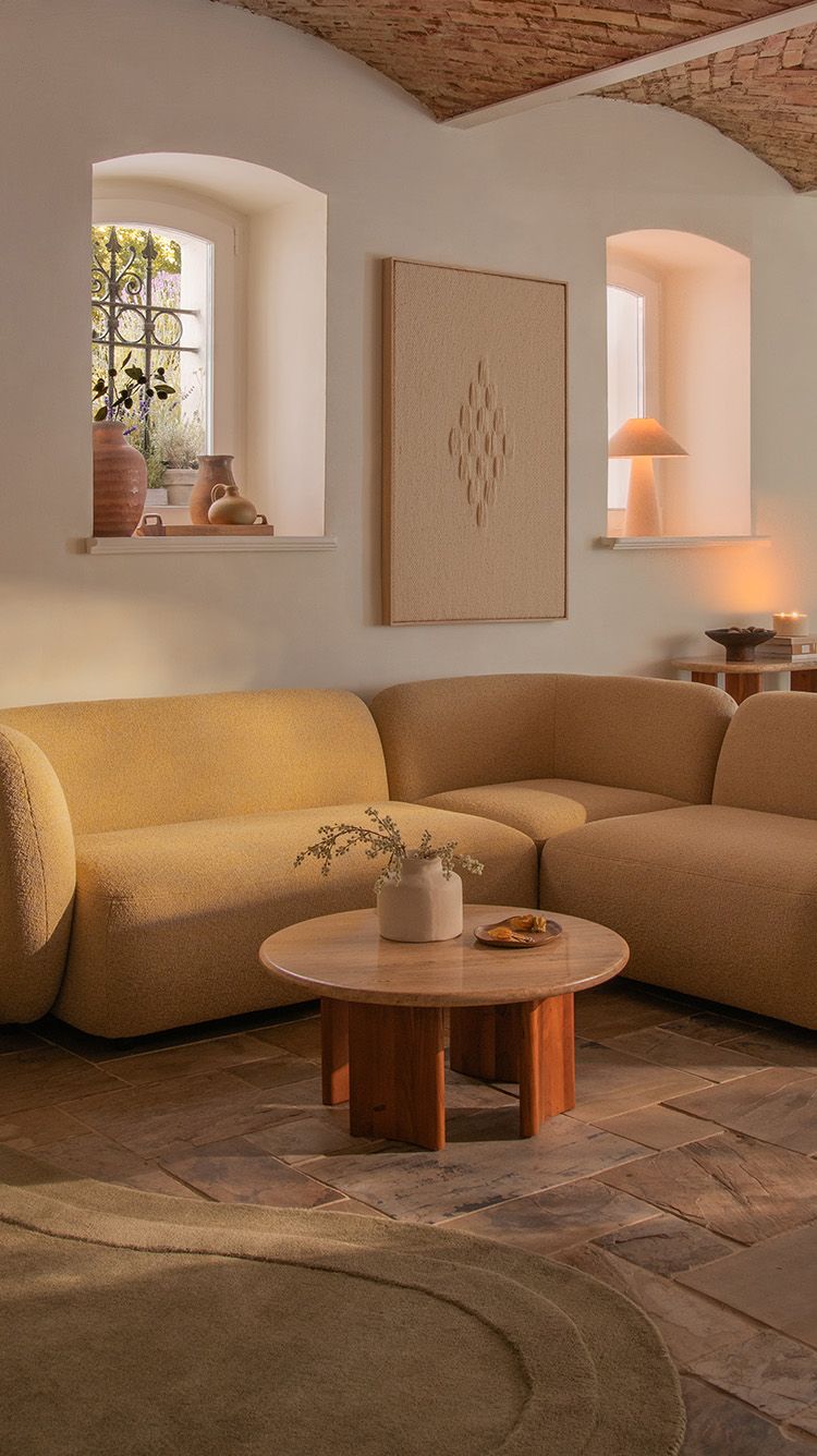 Canapé d’angle Kate en tissu bouclé chaud aux accoudoirs arrondis, associé à la table basse ronde Bia au plateau en pierre et base en bois dans un salon baigné de soleil.