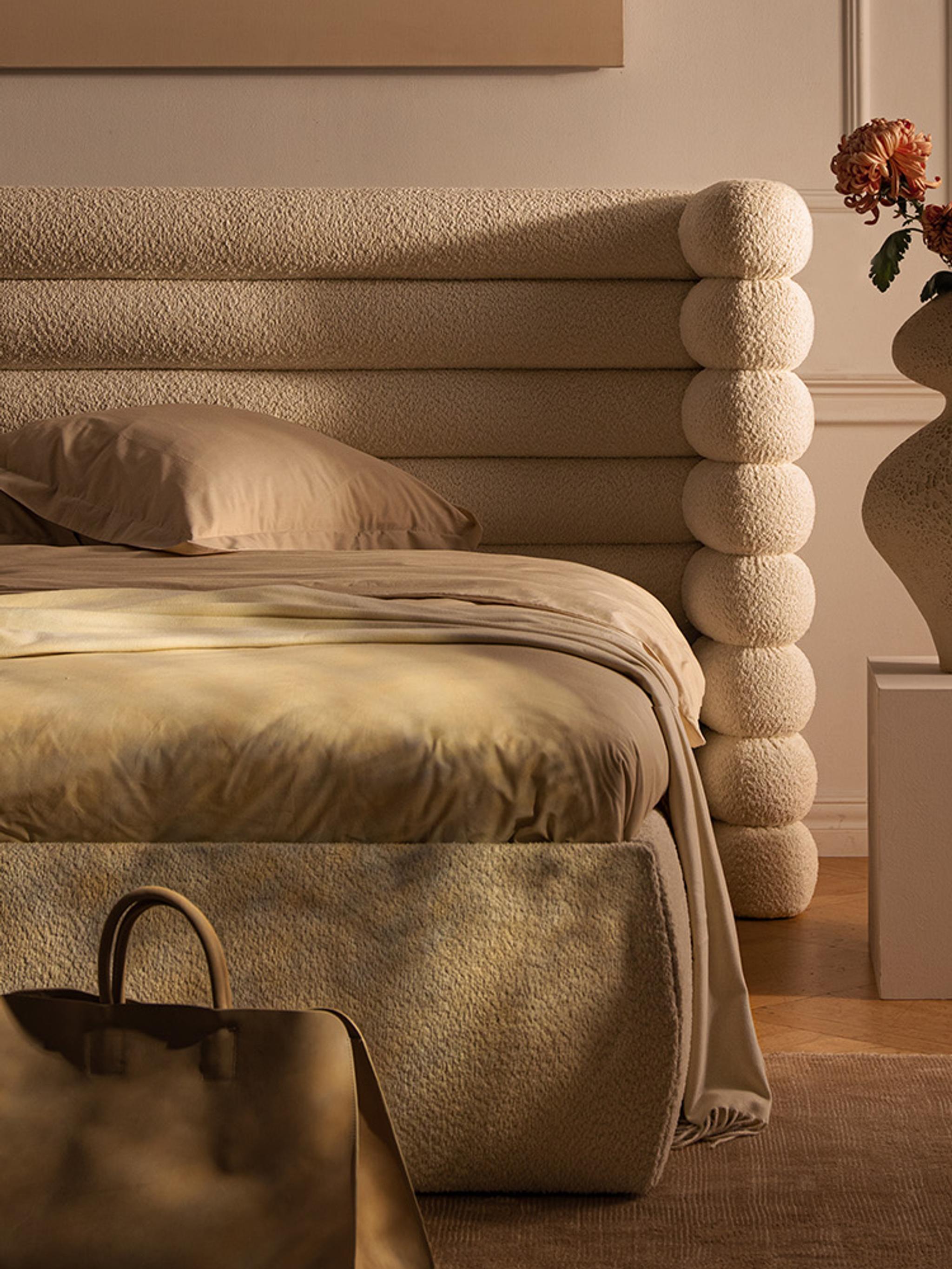 Lit Micadoni Mount avec grande tête de lit à boudins superposés en bouclé crème, paré de linge neutre, posé sur un tapis dans une chambre chaleureuse.