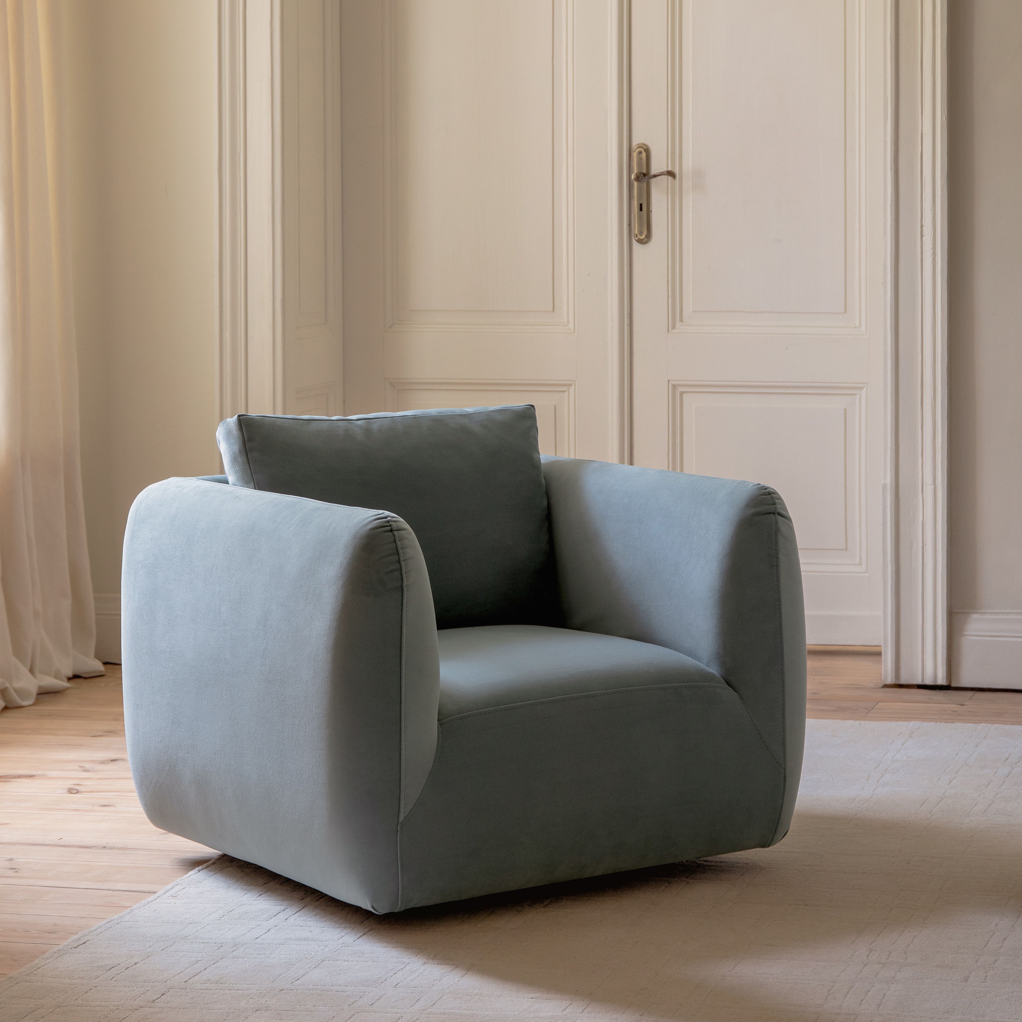 Fauteuil Micadoni Iga avec base pivotante 360° dissimulée, tissu bleu doux et accoudoirs arrondis, posé sur un tapis clair.
