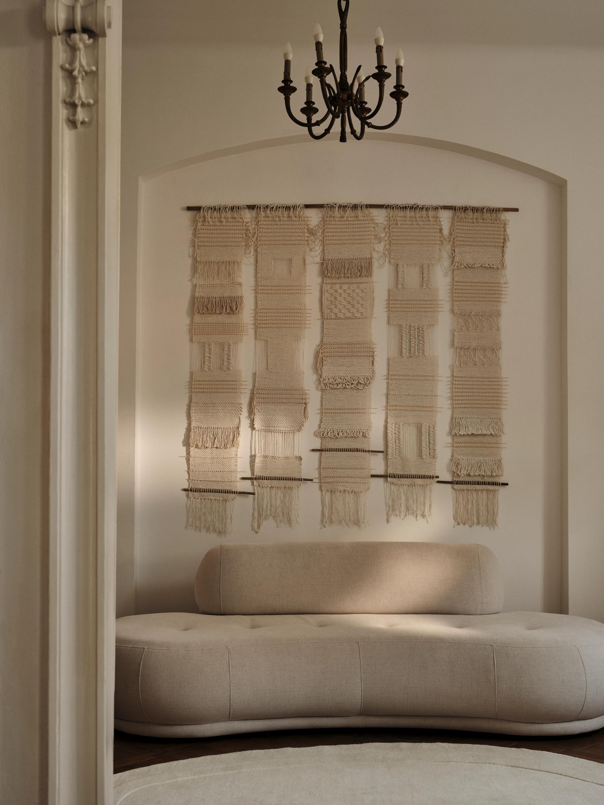Un intérieur chaleureux et élégant mettant en valeur le canapé arrondi Flow de Micadoni, revêtu d’un tissu beige doux. Sa forme organique s’intègre harmonieusement dans un décor minimaliste. Au-dessus, une grande tenture murale en macramé faite à la main ajoute de la texture et de la chaleur. Un lustre en fer forgé noir vient parfaire l’ambiance sophistiquée.