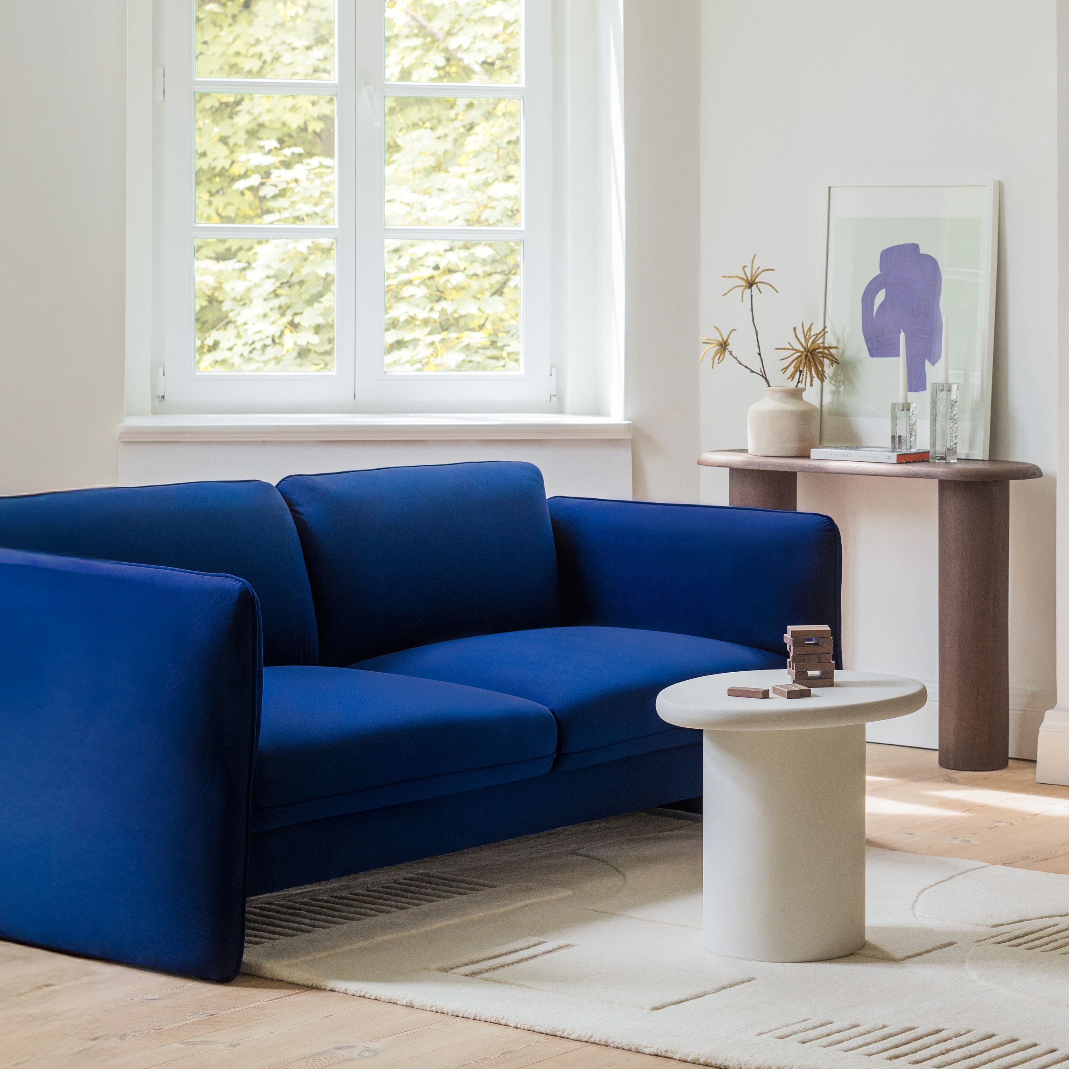 Salon lumineux avec canapé Micadoni Georgia et table basse piédestal ronde blanche Micadoni Daria sur tapis crème, près d’une fenêtre et d’une console.