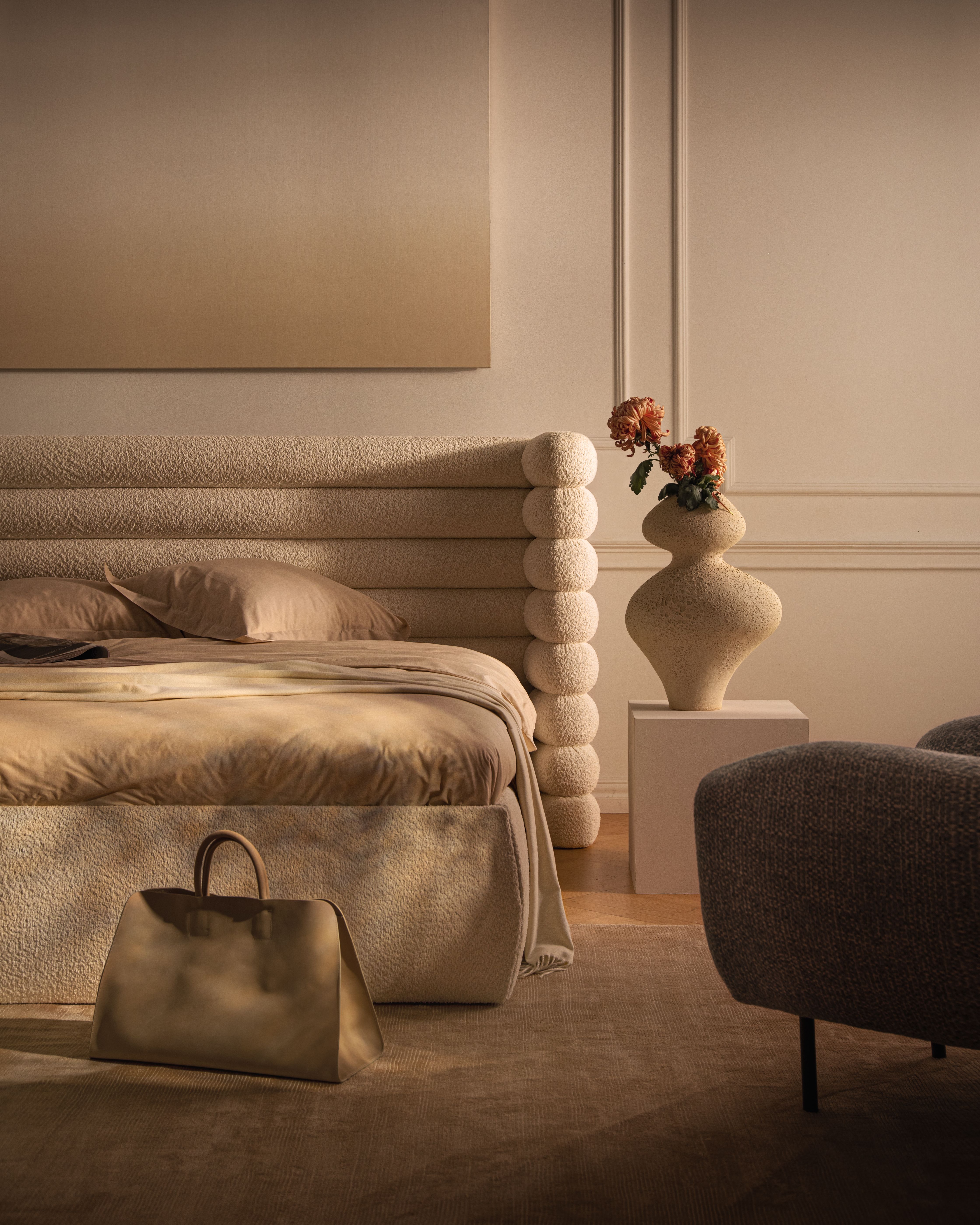 Lit Micadoni en bouclé avec tête de lit sculpturale en couches, linge beige, vase sur socle, lumière chaude.