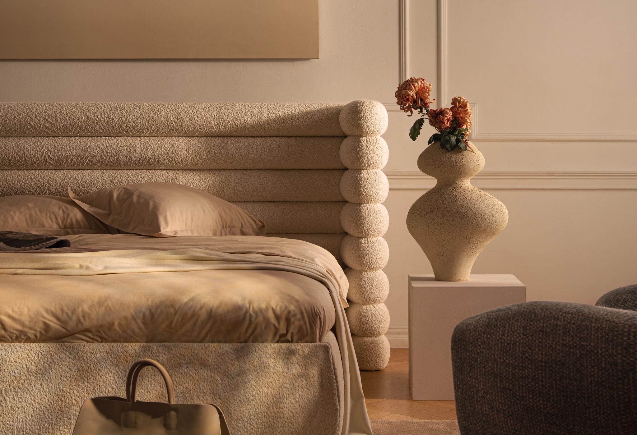 Lit Micadoni en bouclé avec tête de lit sculpturale en couches, linge beige, vase sur socle, lumière chaude.