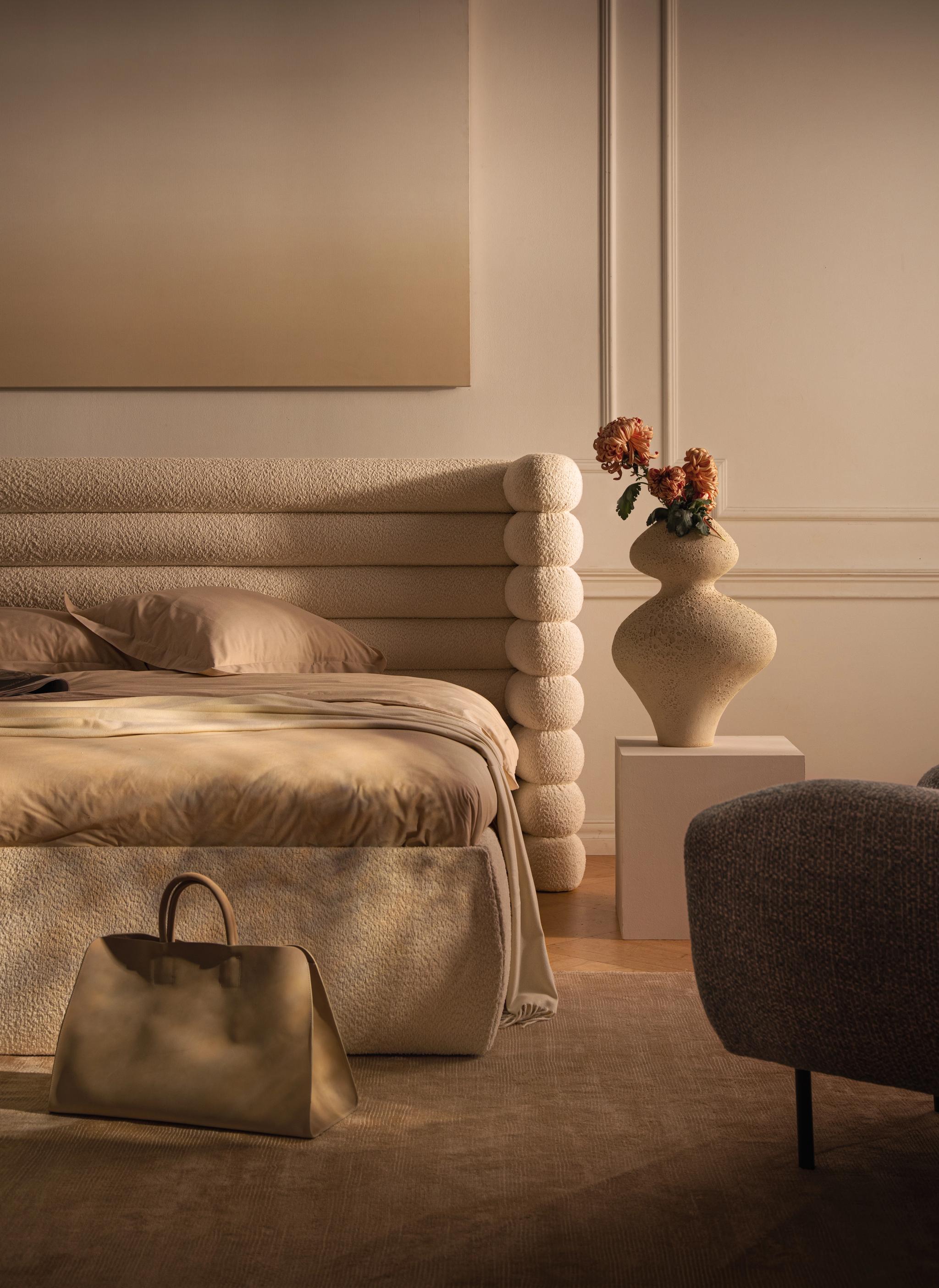 Lit Micadoni en bouclé avec tête de lit sculpturale en couches, linge beige, vase sur socle, lumière chaude.