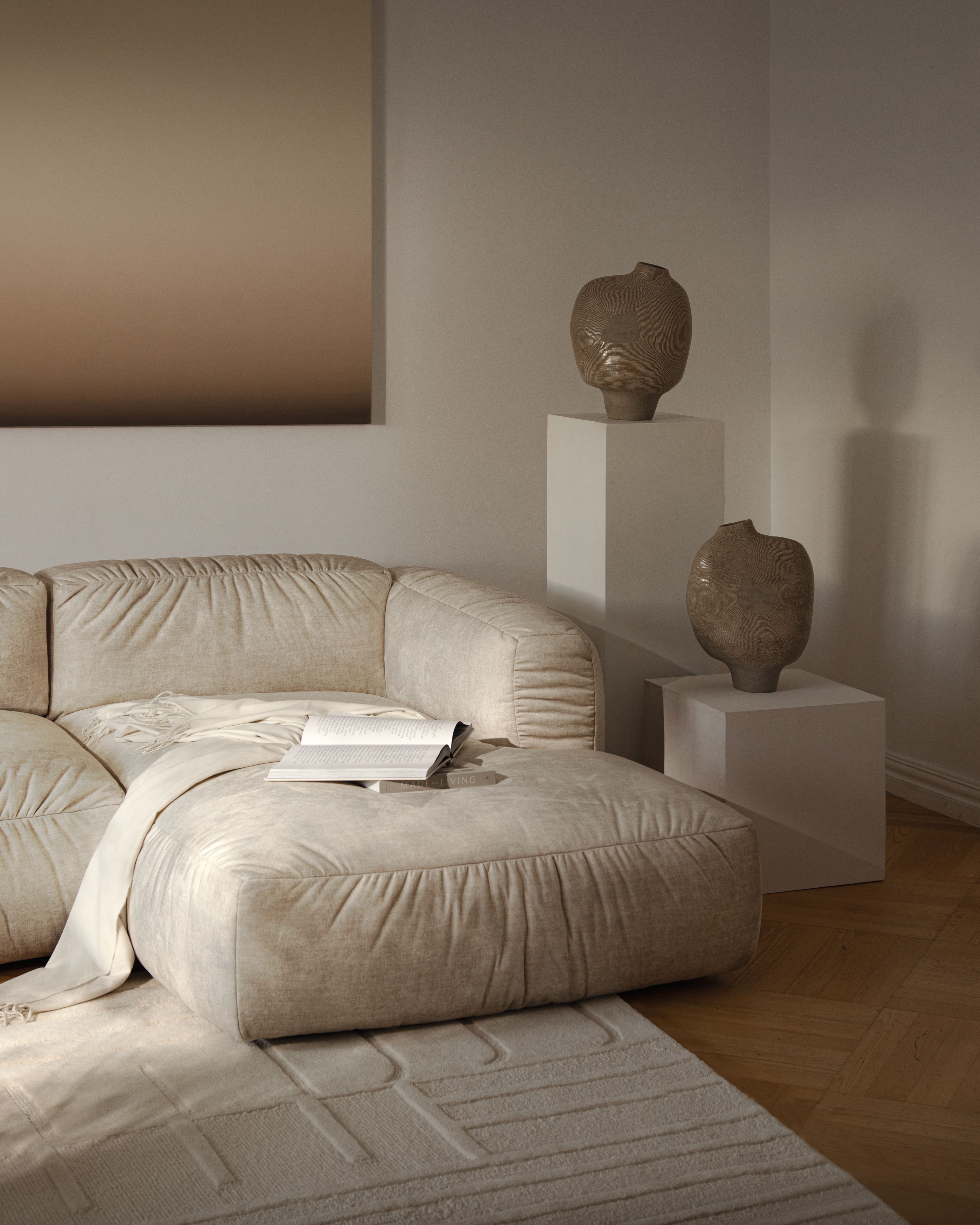 Canapé d’angle Micadoni Martina en tissu beige texturé, coutures froncées et large méridienne, posé sur un tapis à relief avec vases en céramique sur socles, lumière douce.