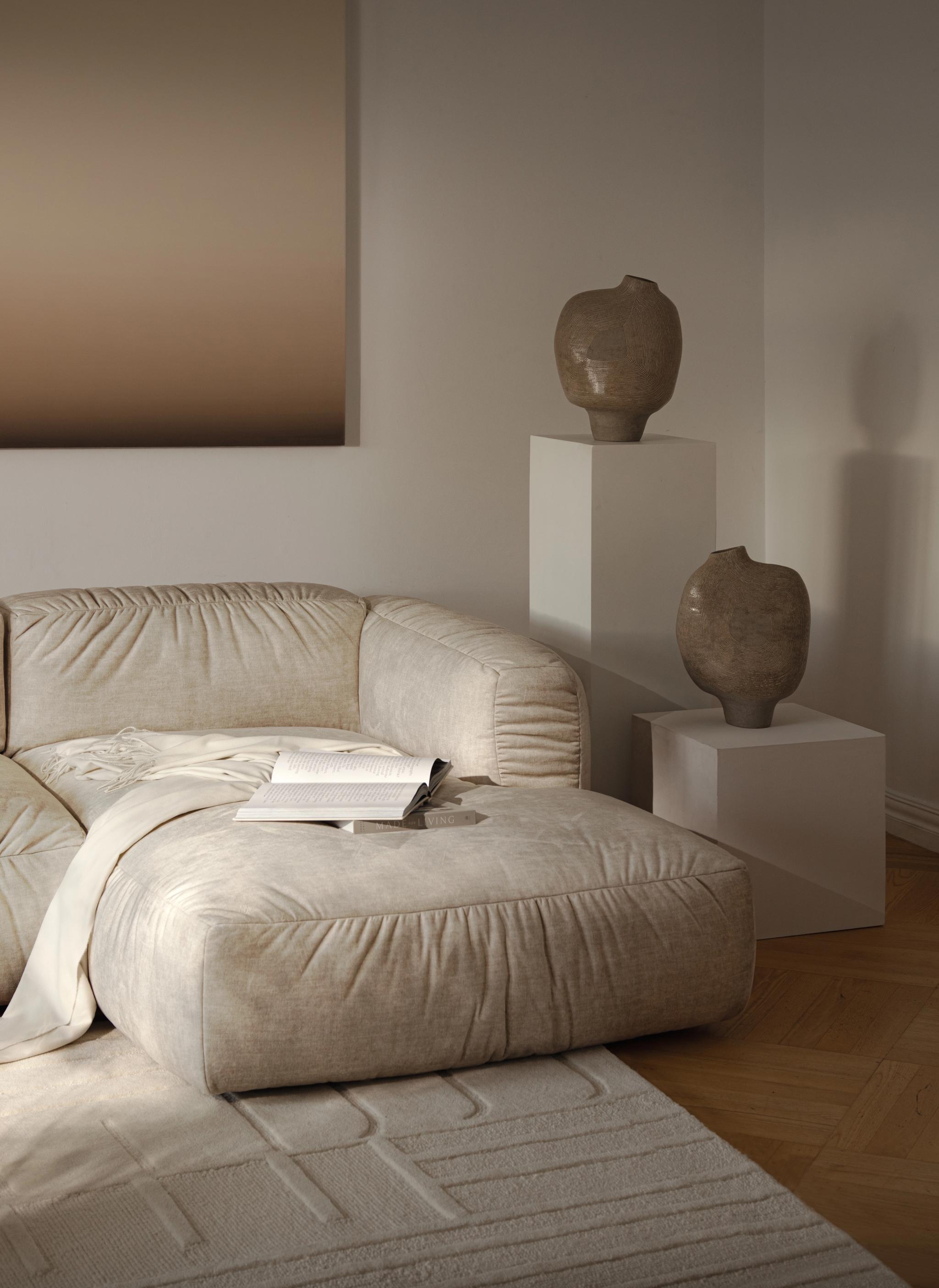 Canapé d’angle Micadoni Martina en tissu beige texturé, coutures froncées et large méridienne, posé sur un tapis à relief avec vases en céramique sur socles, lumière douce.