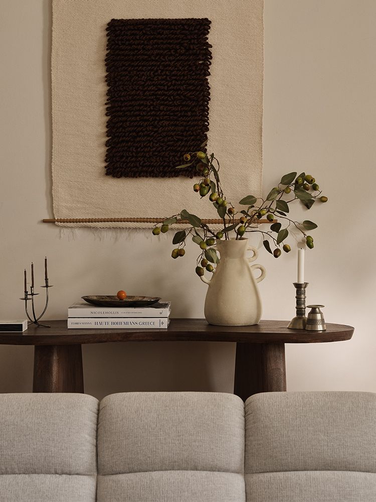 Scène salon Micadoni avec console en bois foncé, pichet en céramique et branches, livres et bougie, tenture murale textile et dossier d’un canapé gris clair.