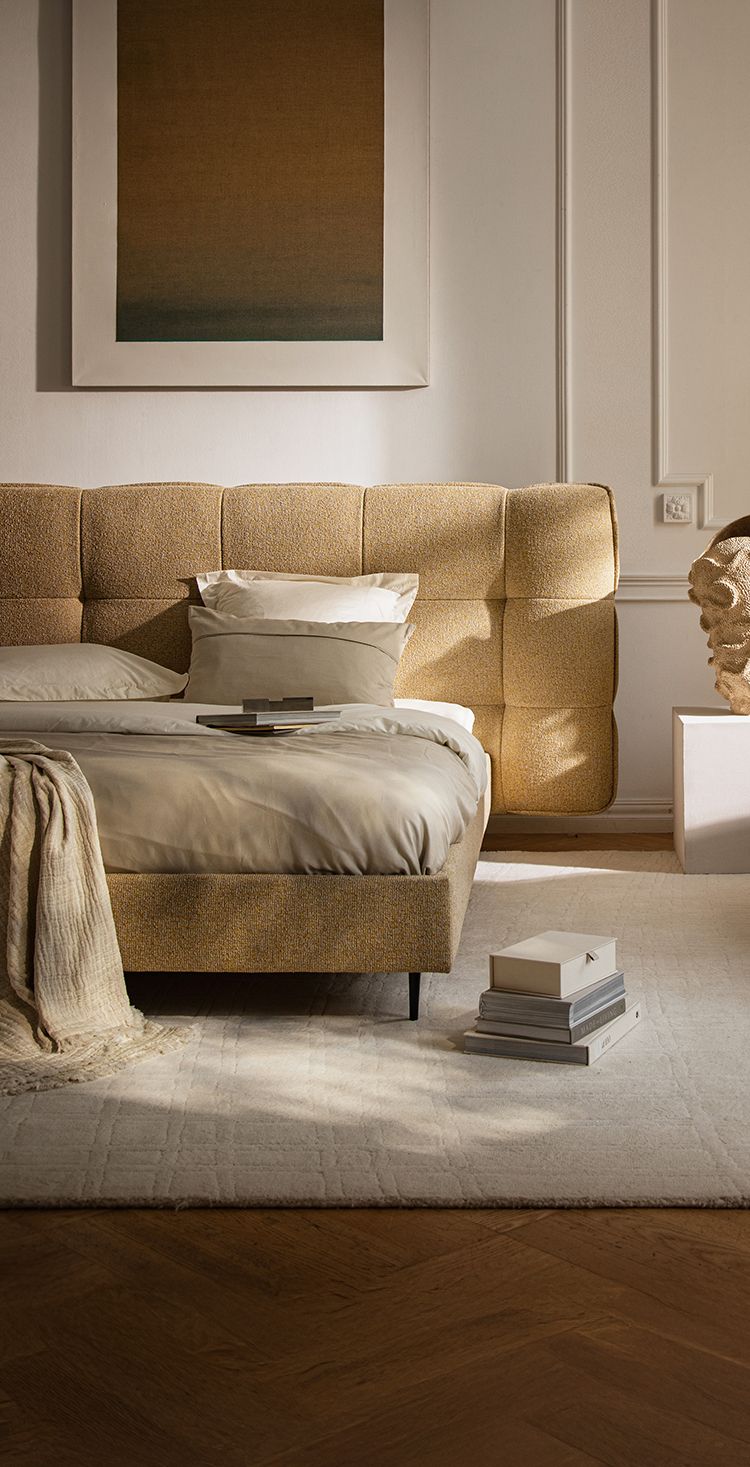 Lit Micadoni avec tête de lit capitonnée arrondie et fauteuil gris texturé dans une chambre chaleureuse.