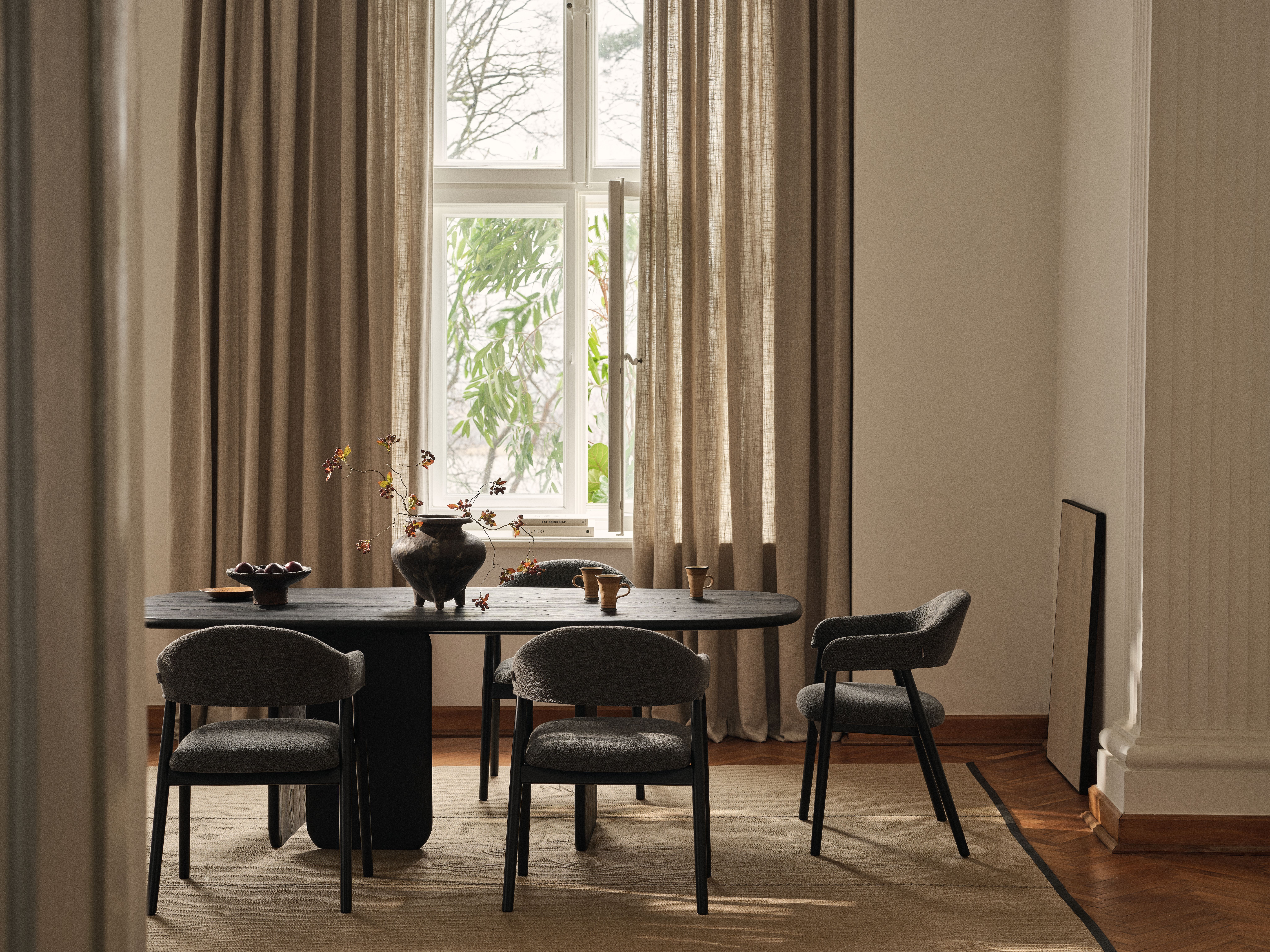 Salle à manger moderne Micadoni avec table ovale noire et quatre chaises capitonnées sombres, rideaux neutres, lumière du jour.
