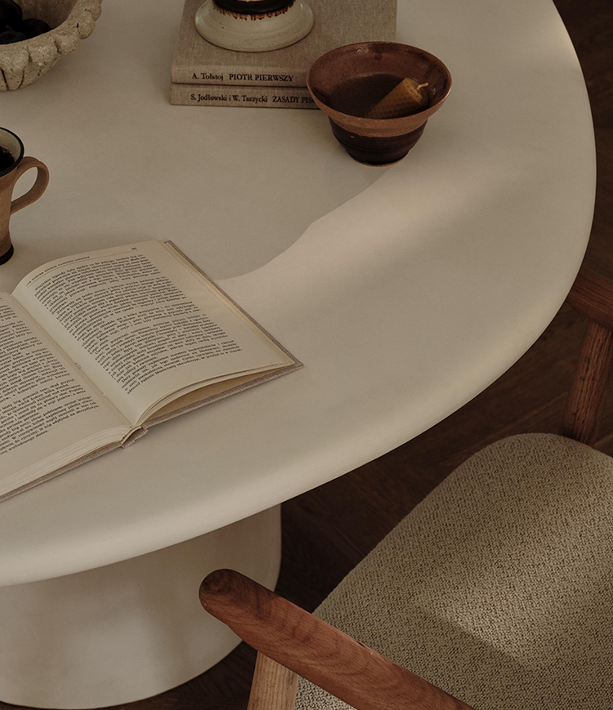 Table de salle à manger Micadoni Daria dans une ambiance chaleureuse, avec céramiques, livre ouvert et fauteuil texturé.